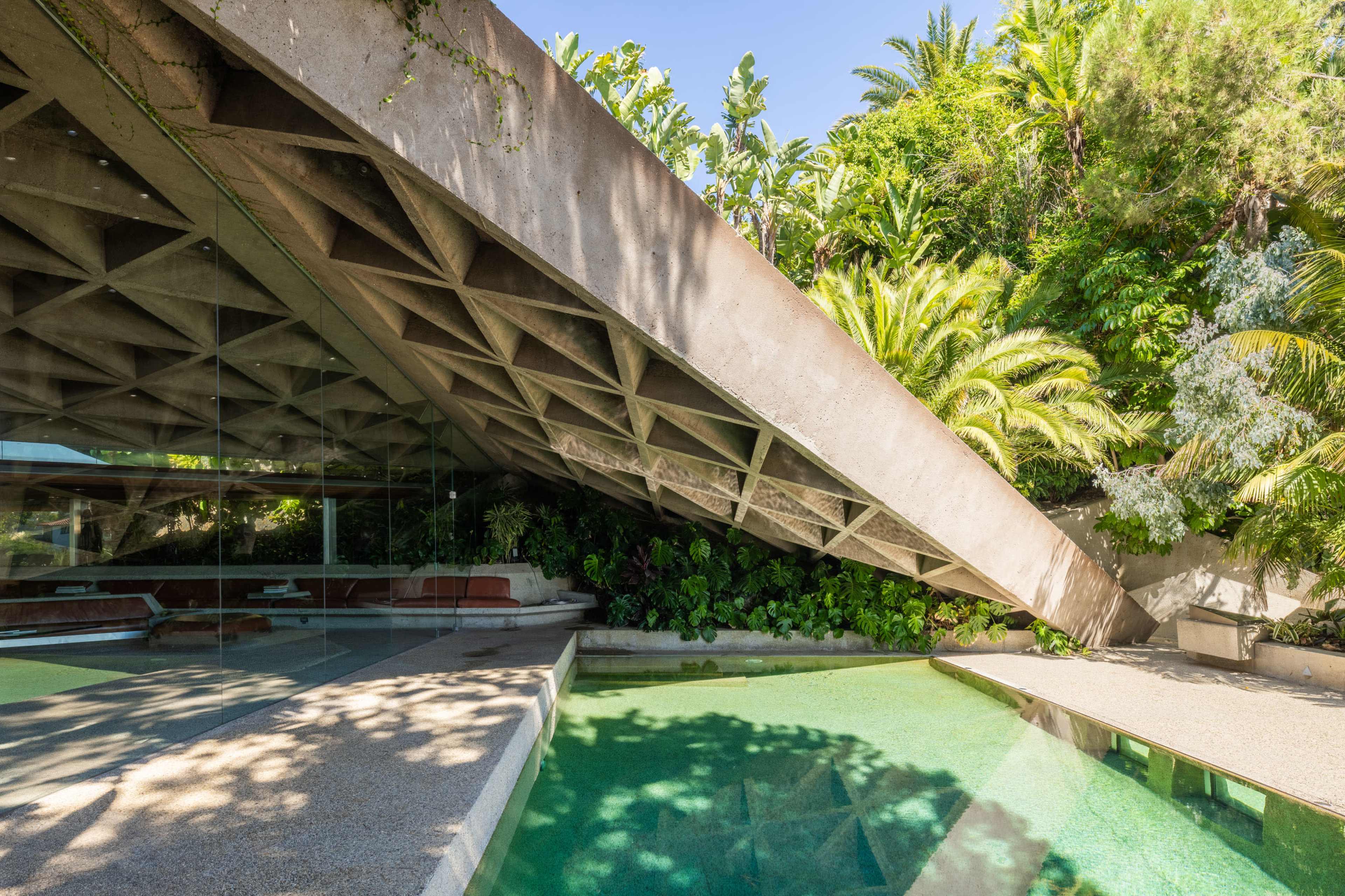A modern architectural structure with a distinctive concrete roof extending over a shallow pool surrounded by tropical foliage.