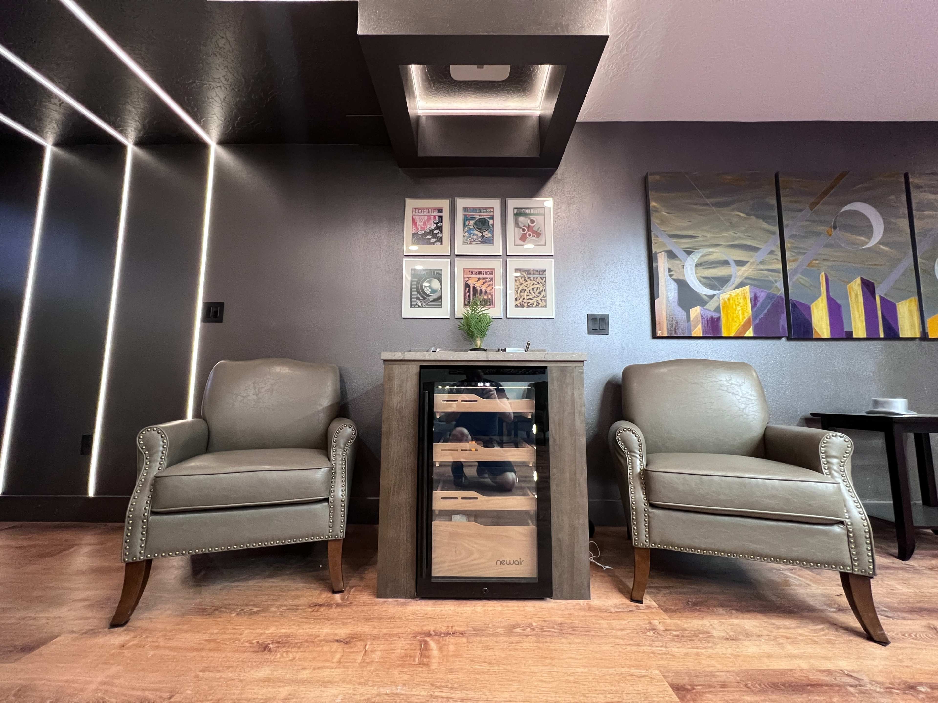 Two gray chairs flanking a modern wine cooler on a wooden floor, with framed artwork on the wall behind.