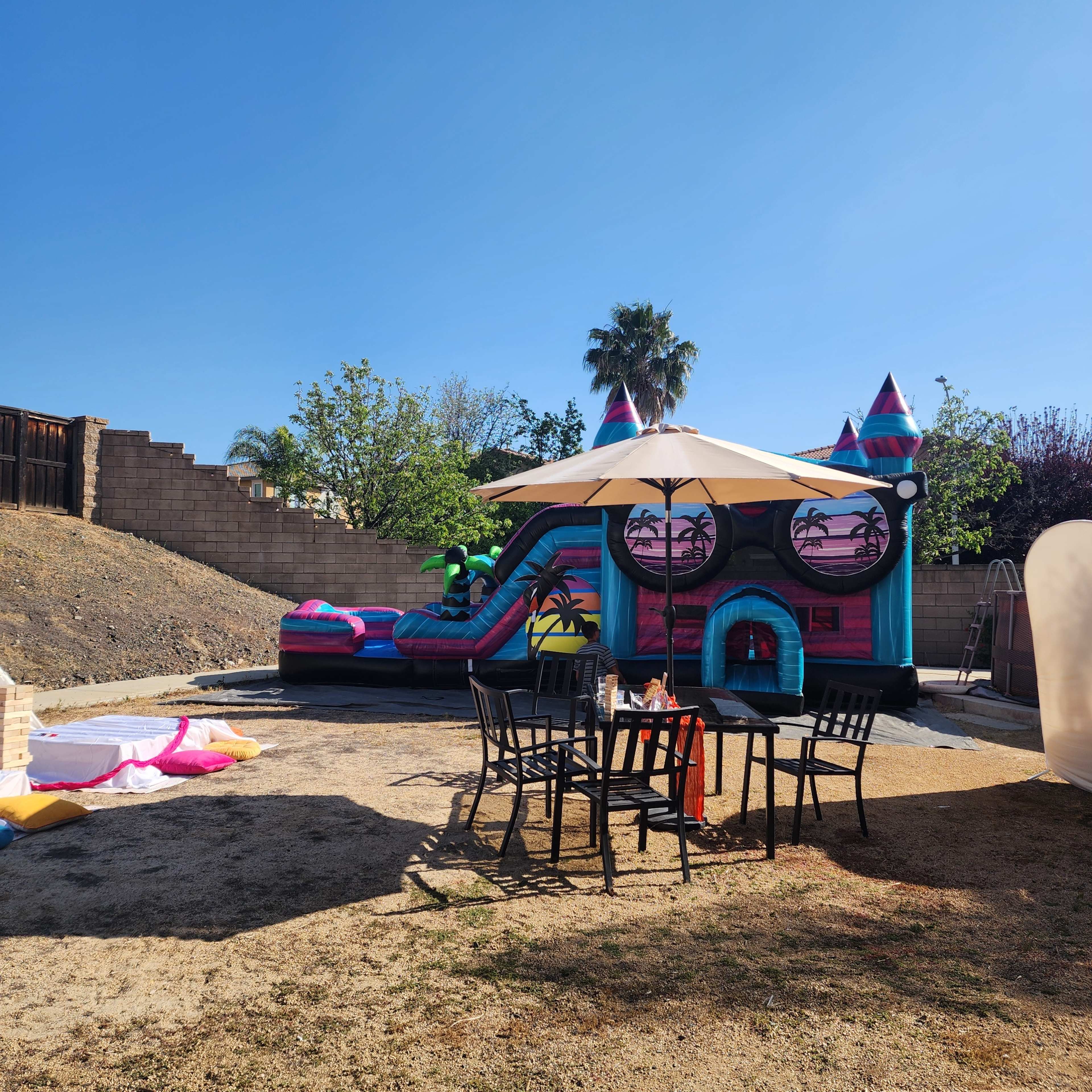 A colorful inflatable bounce house with a water slide is set up in a sandy backyard, accompanied by a table and chairs beneath an umbrella.