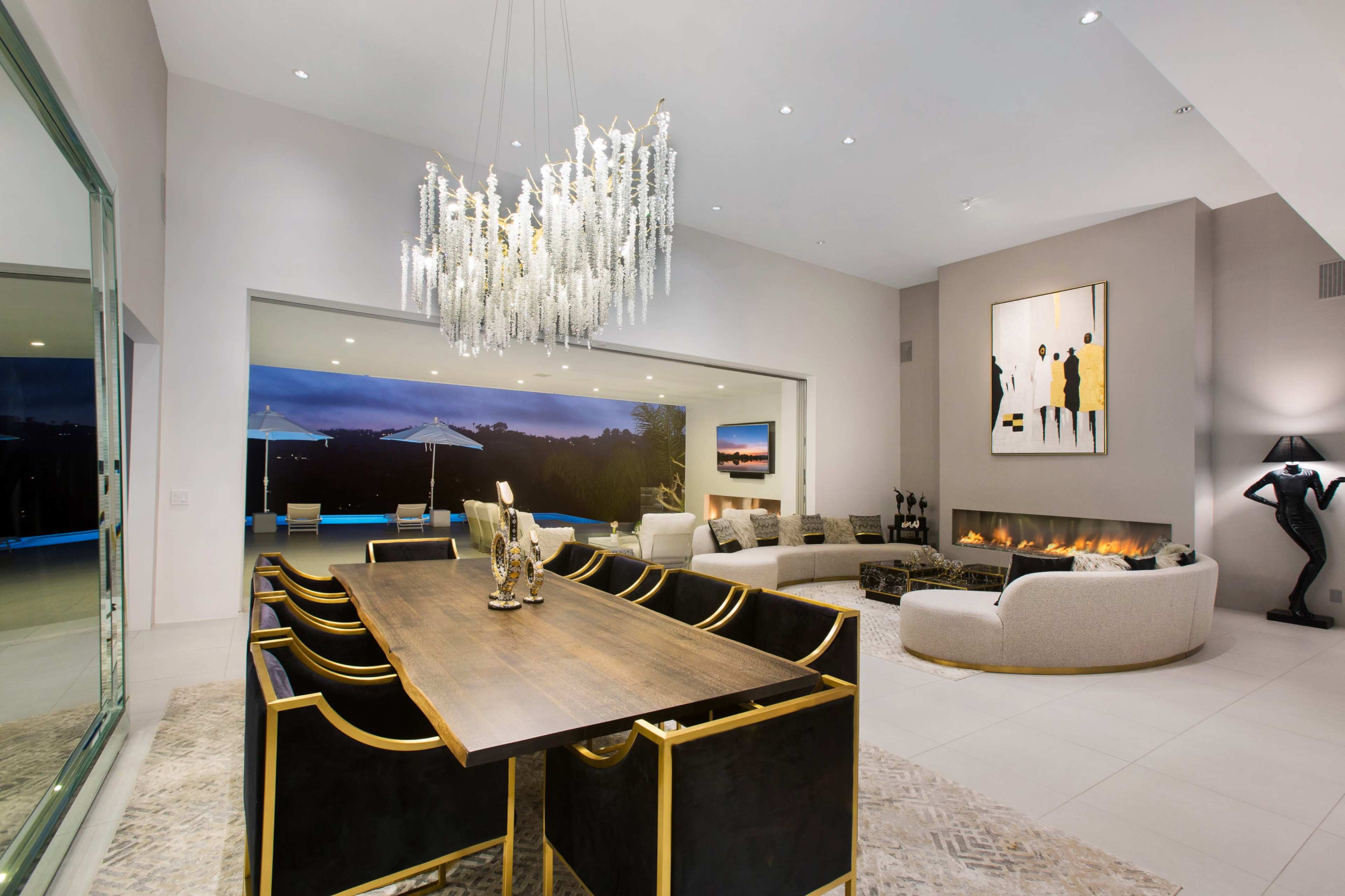 The image depicts a modern living room space featuring a large dining table, a curved sofa, a striking chandelier, and a fireplace, with large glass doors leading to an outdoor area.