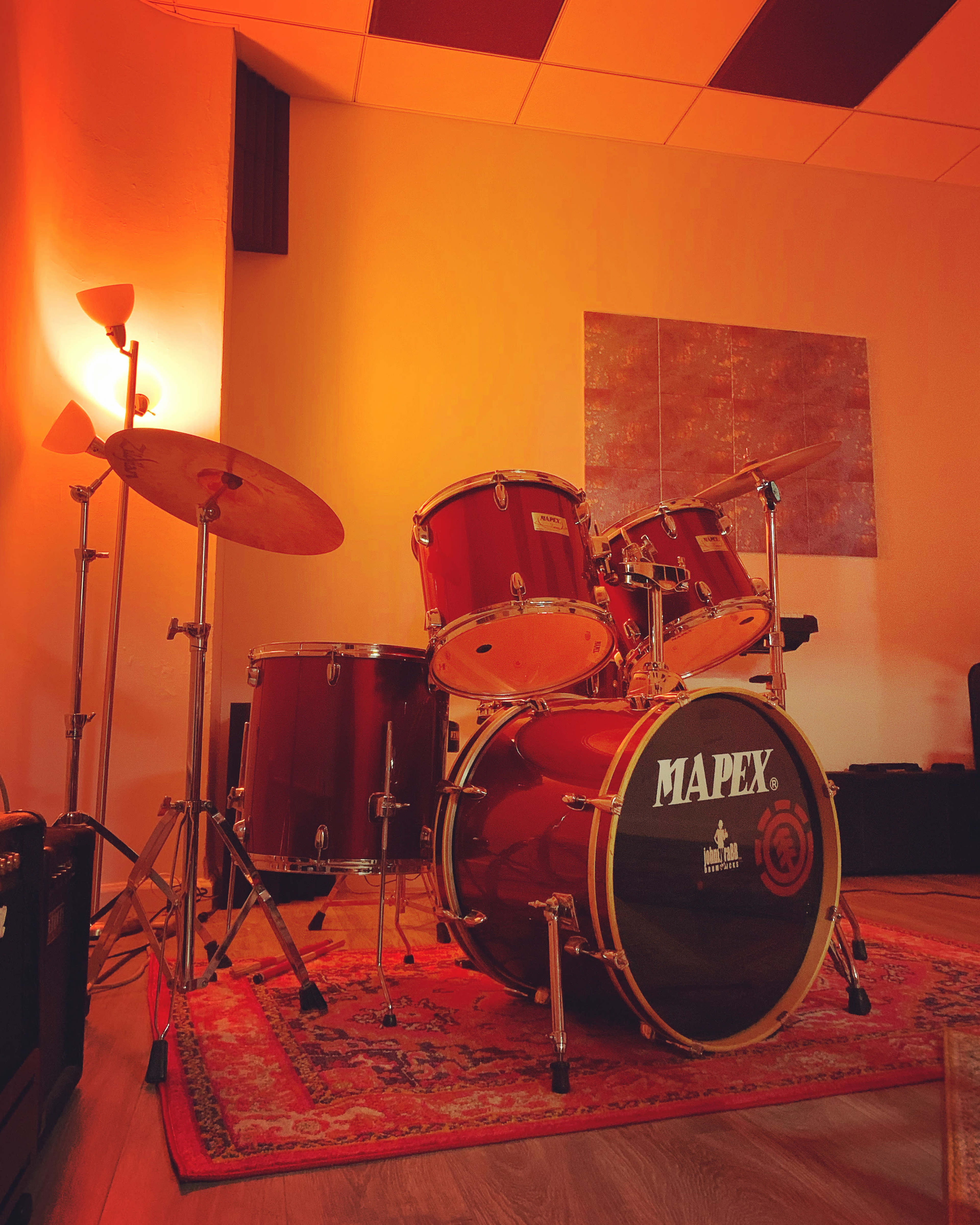 A red drum kit is positioned on a patterned rug in a warmly lit room with a lamp and a wall art piece.