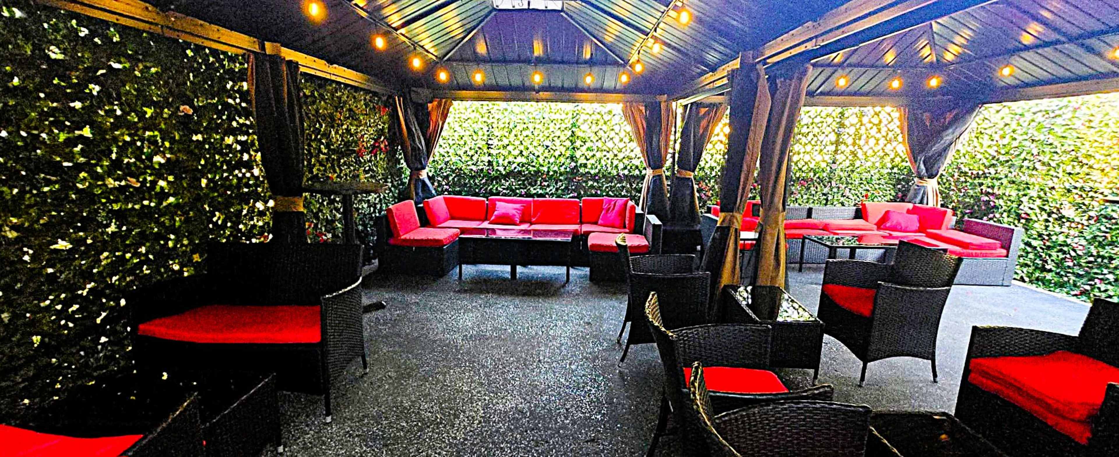 The image shows a covered outdoor patio area with red seating and surrounding greenery, illuminated by string lights.