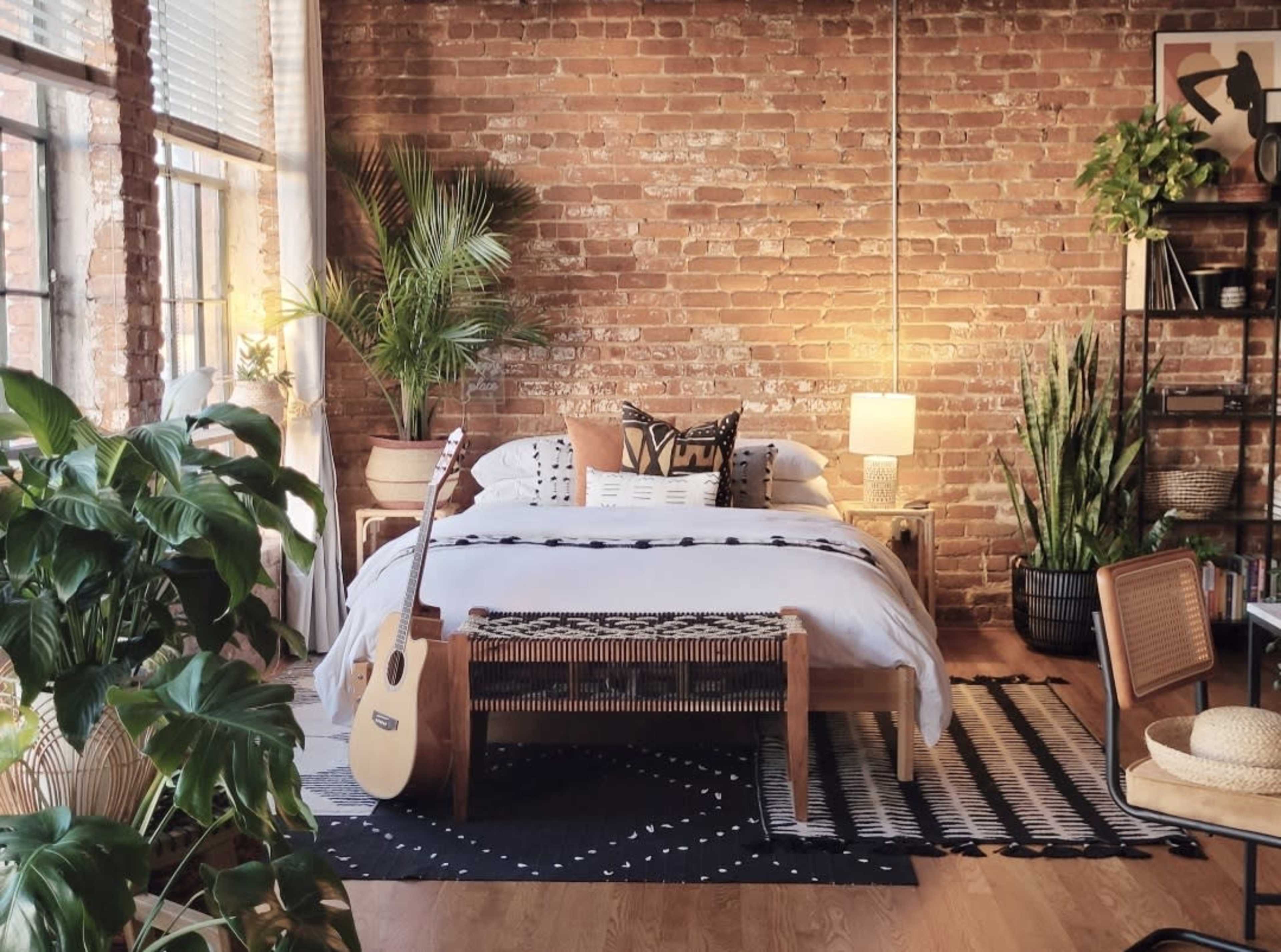 A cozy bedroom with a brick wall, a bed adorned with decorative pillows, and a guitar and plants arranged around the space.