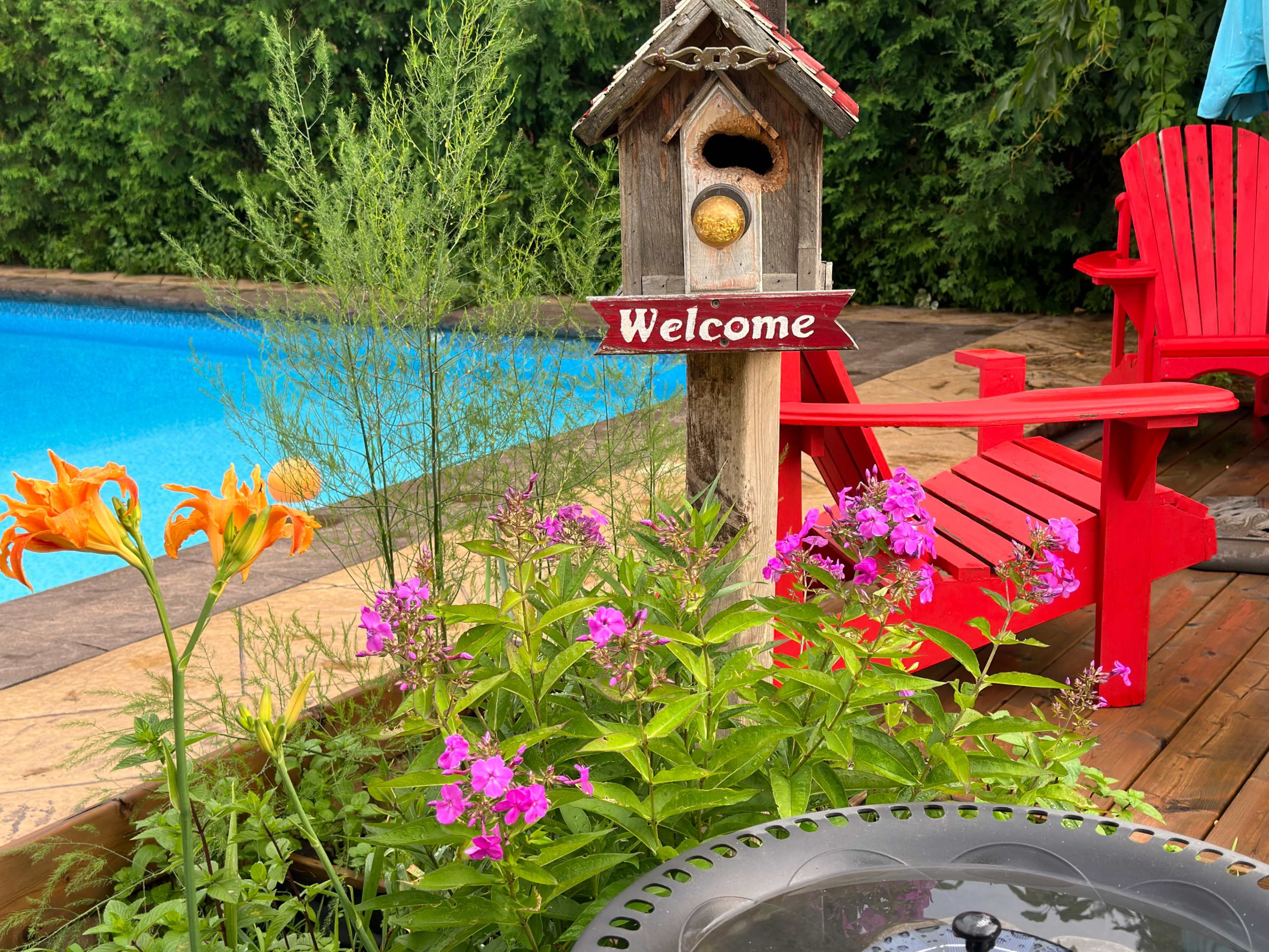 A wooden birdhouse with a "Welcome" sign stands beside colorful flowers and red adirondack chairs near a swimming pool.