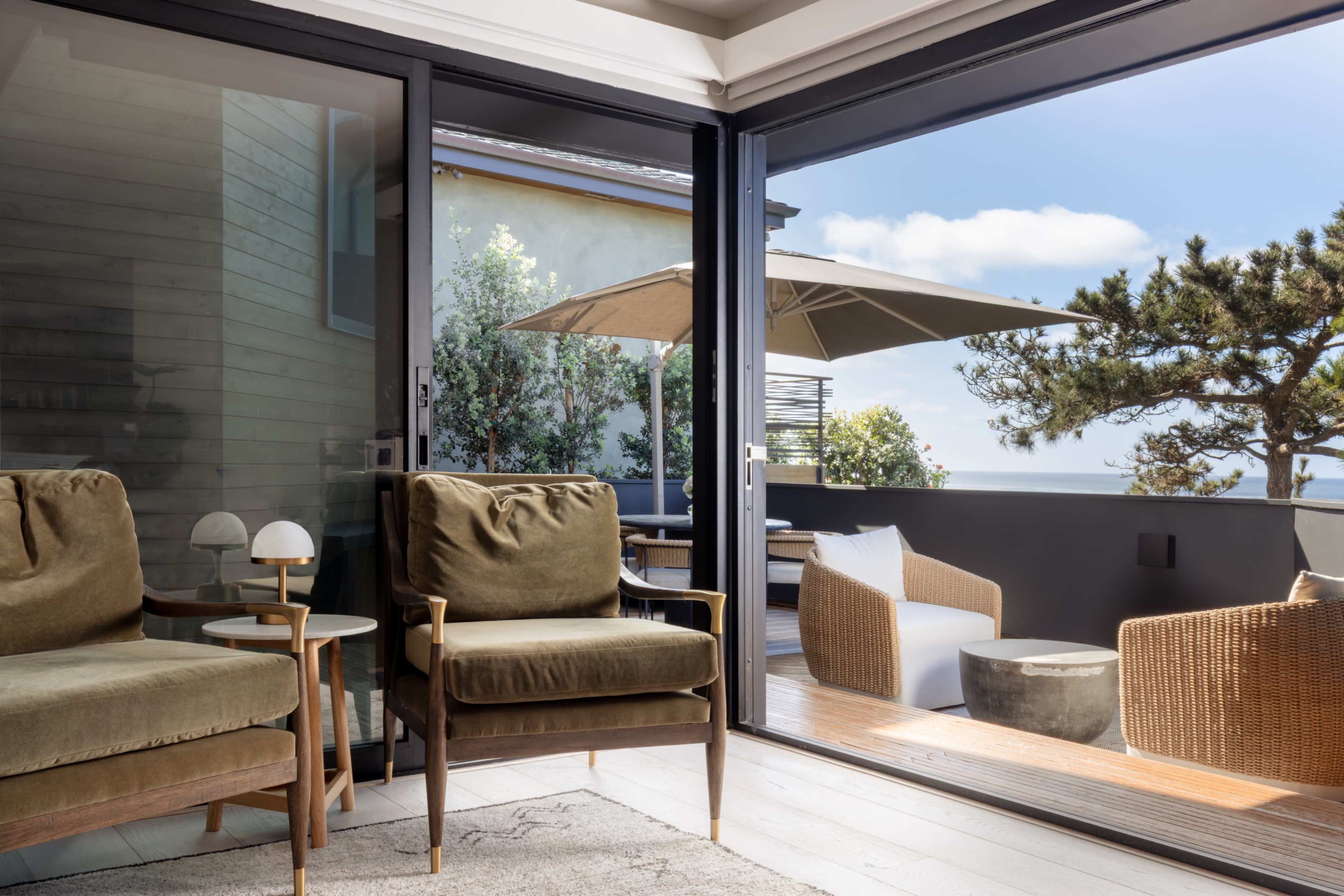 The image shows a modern living space with two upholstered chairs facing a glass door that opens to a patio featuring an outdoor seating area and an umbrella.