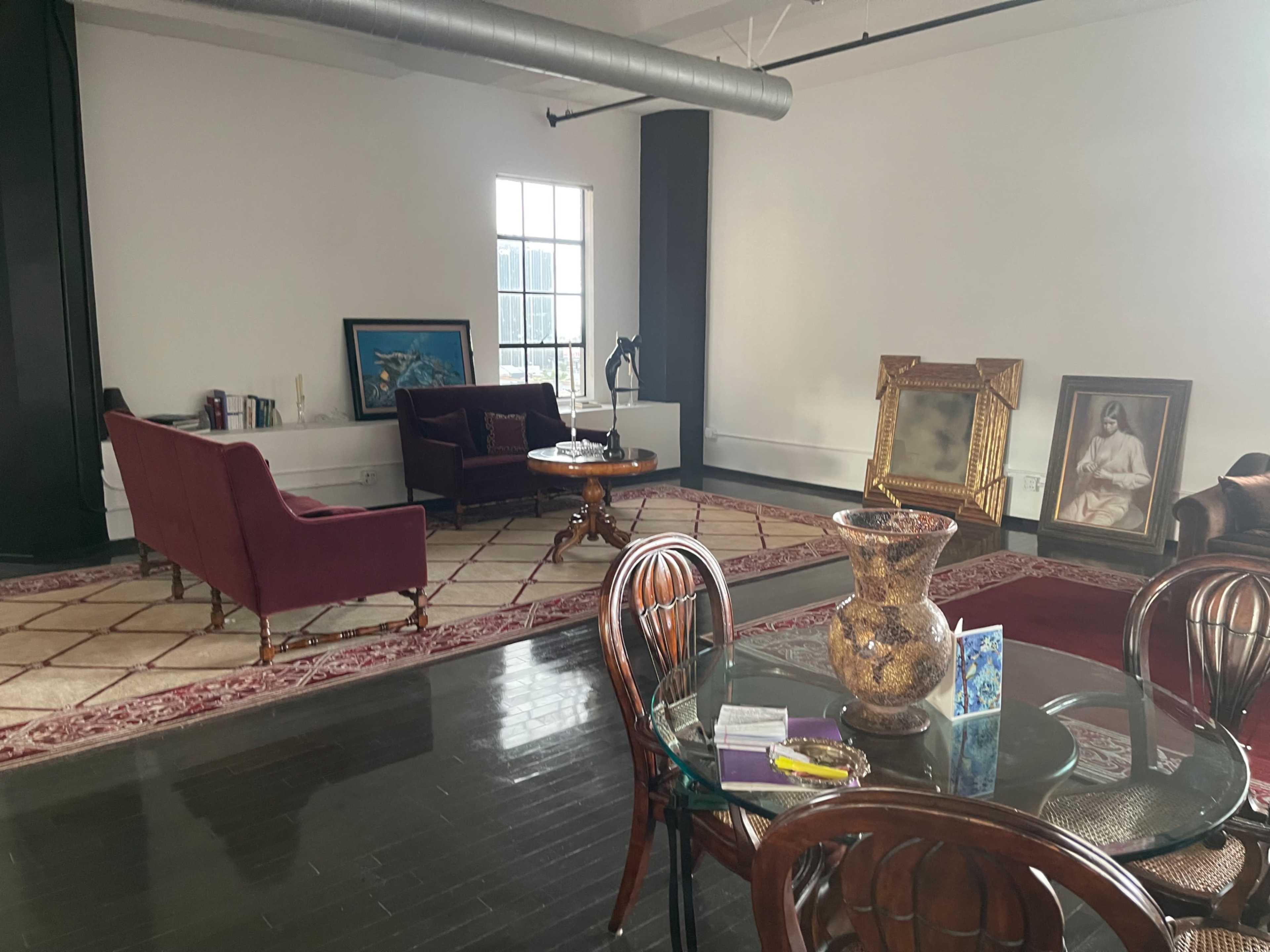 The image shows a spacious, well-lit room with a cozy seating area, featuring a purple couch, a wooden coffee table, and several artworks displayed on the walls.