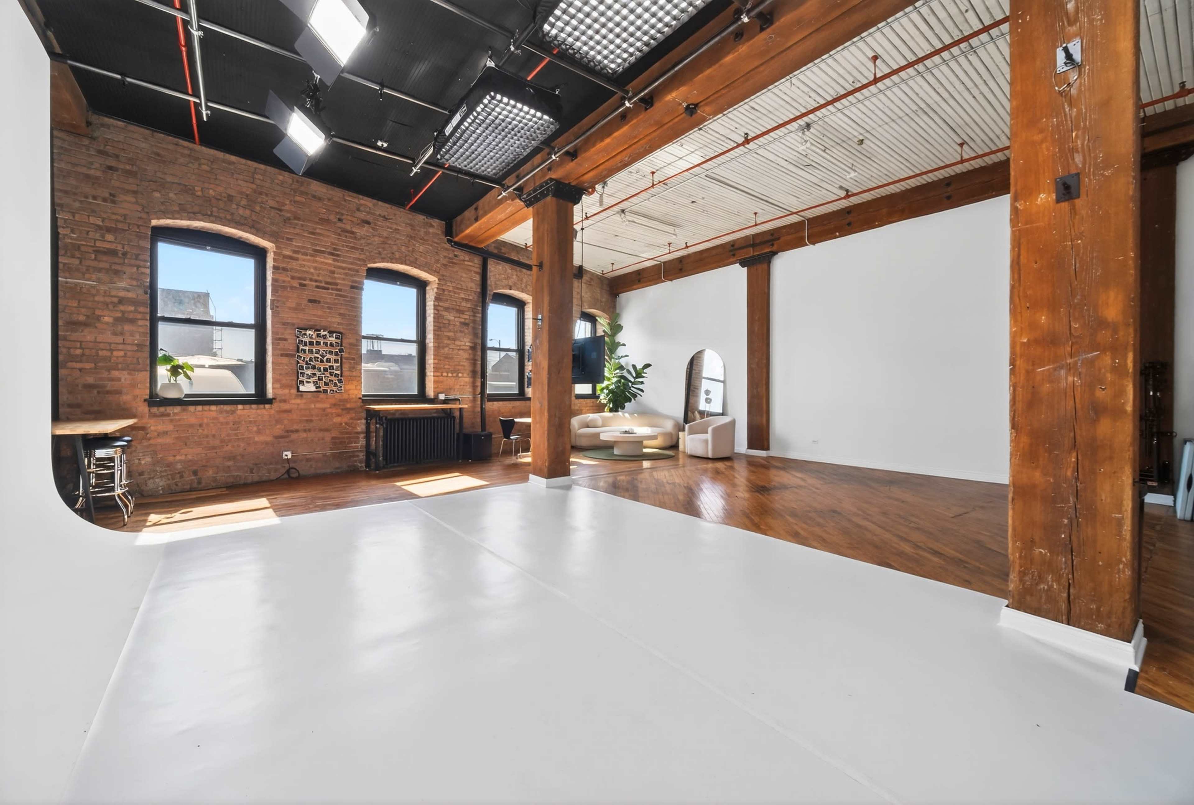 The image shows a spacious studio with a polished white floor, large windows, exposed brick walls, and wooden beams providing a backdrop for creative activities.