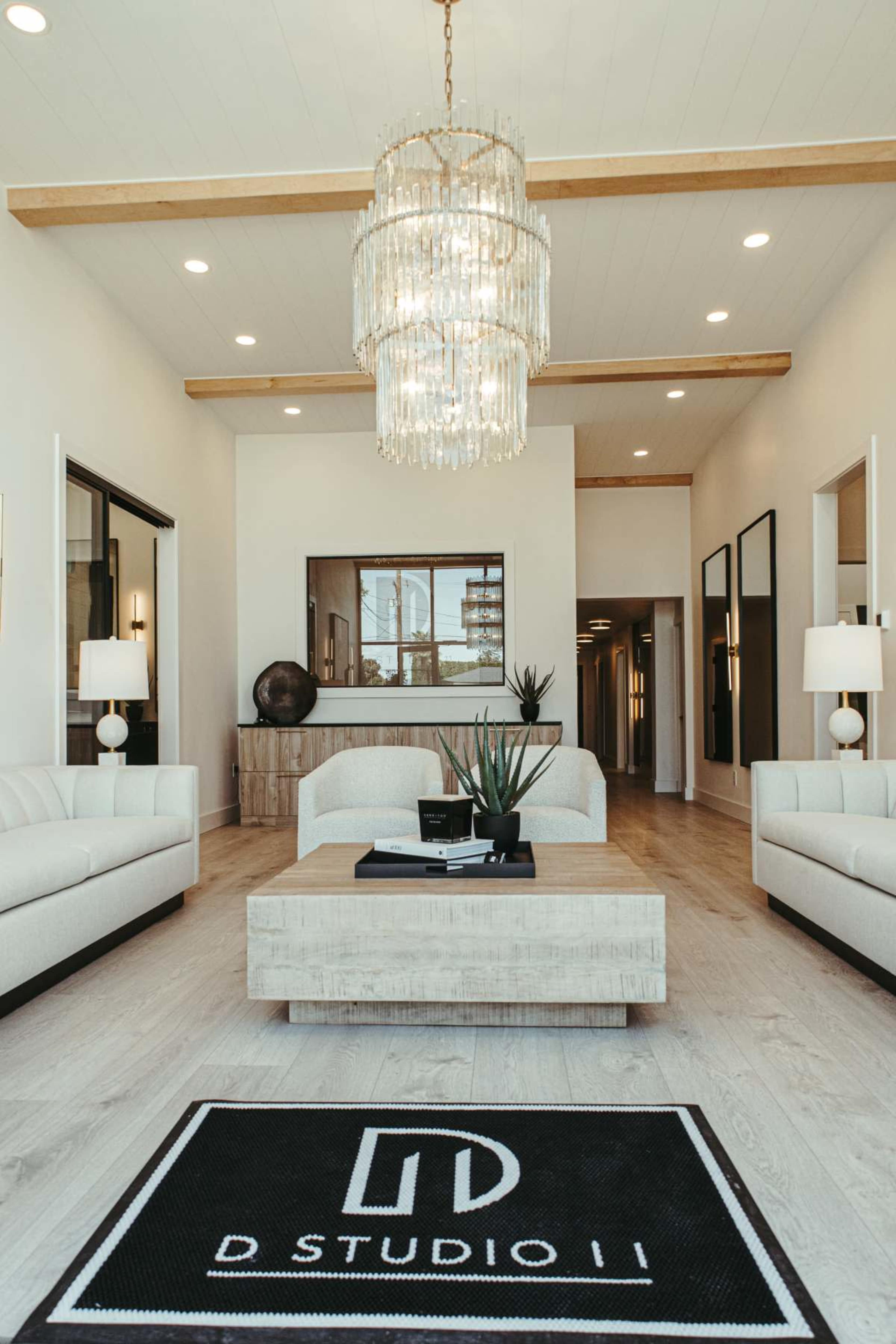 The image shows a modern interior of a lobby or waiting area featuring two white sofas, a central wooden coffee table, a large chandelier, and a decorative rug with "D STUDIO II" printed on it.