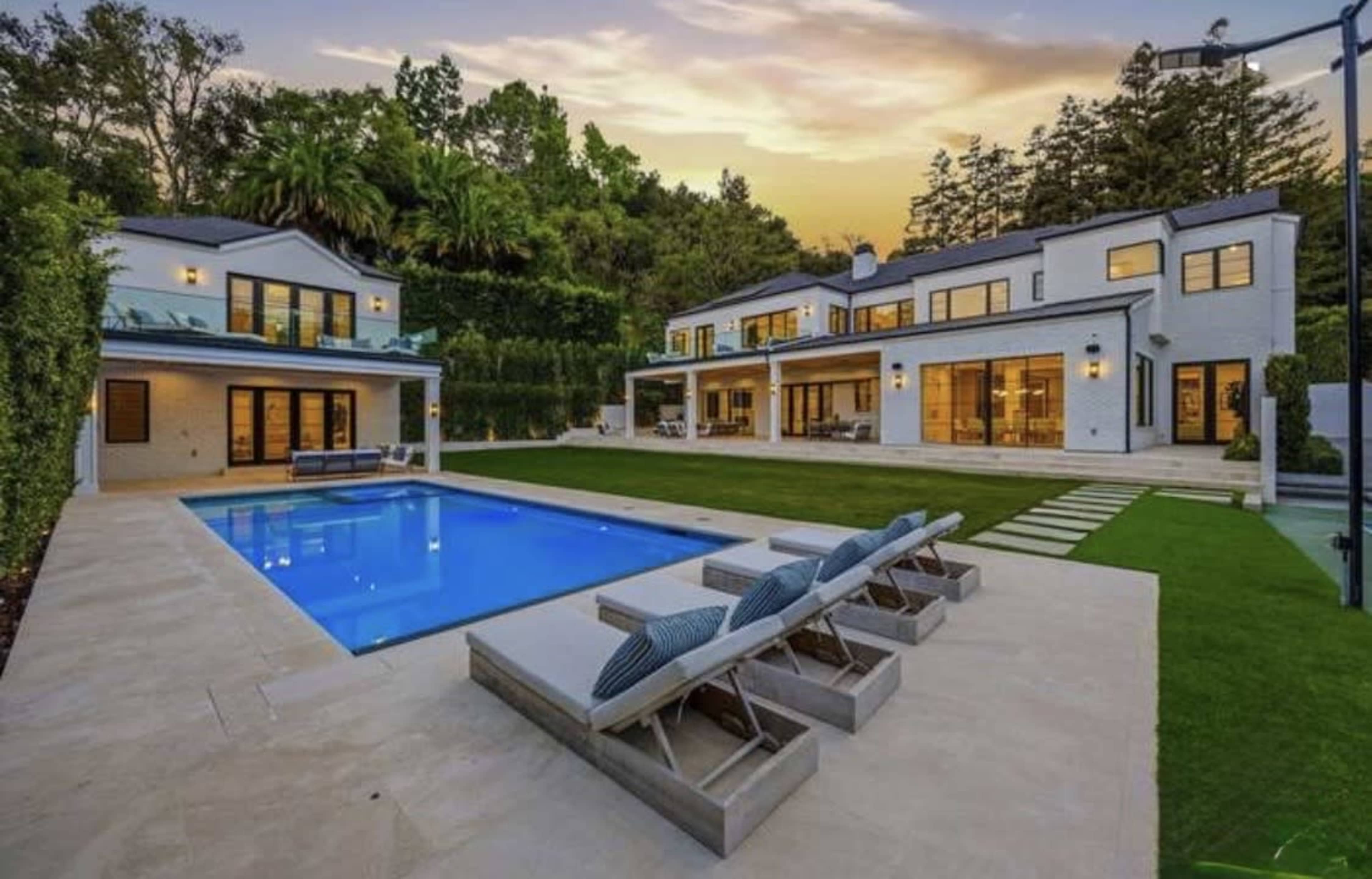 The image shows a modern two-story house with a swimming pool and lounge chairs in a landscaped backyard during sunset.