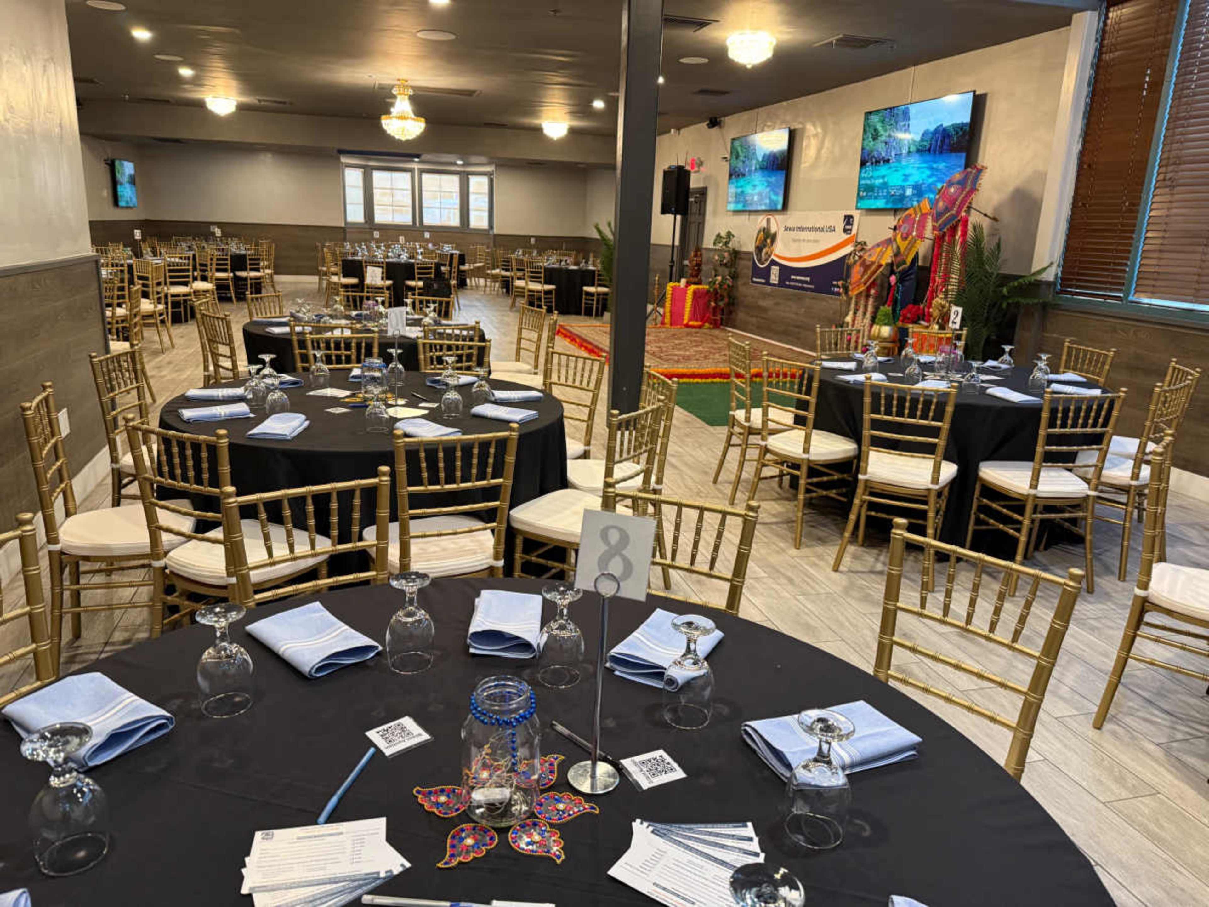 The image shows a well-arranged dining hall with black tablecloths, gold chairs, and large screens displaying colorful images.