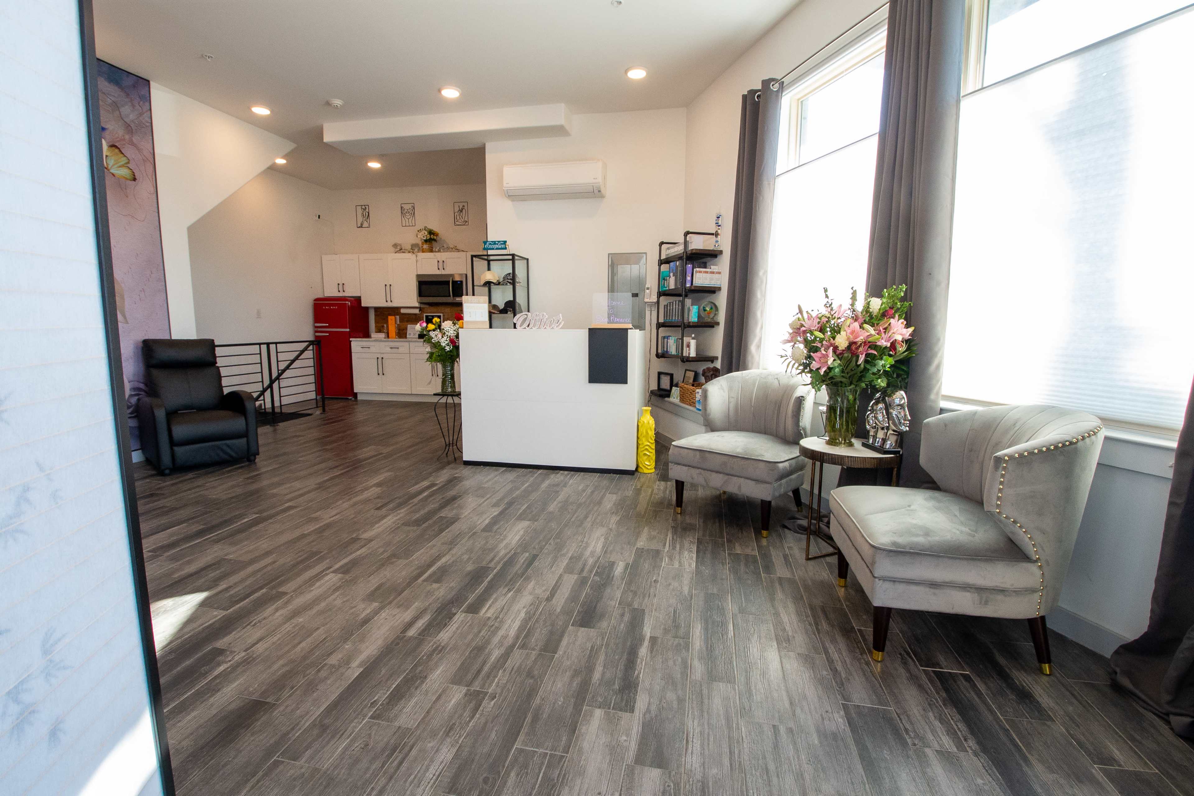 A modern reception area with gray wood flooring, two elegant chairs, a small round table, a white front desk, and a kitchenette in the background.