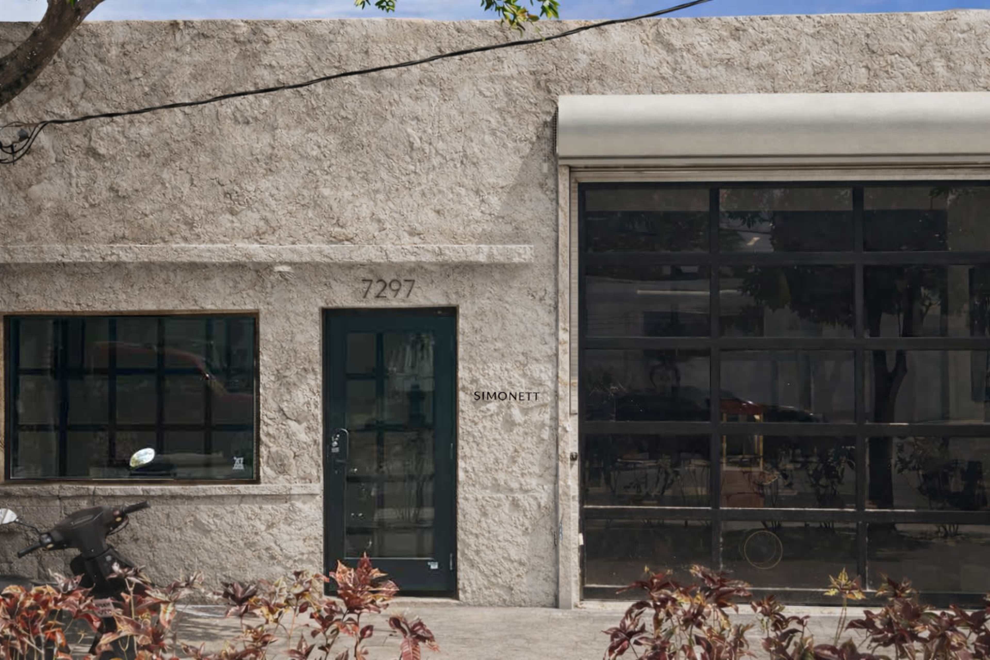 The image shows a textured, light-colored building facade with a green door and a glass garage door, featuring the name "SIMONETT" displayed on the wall.