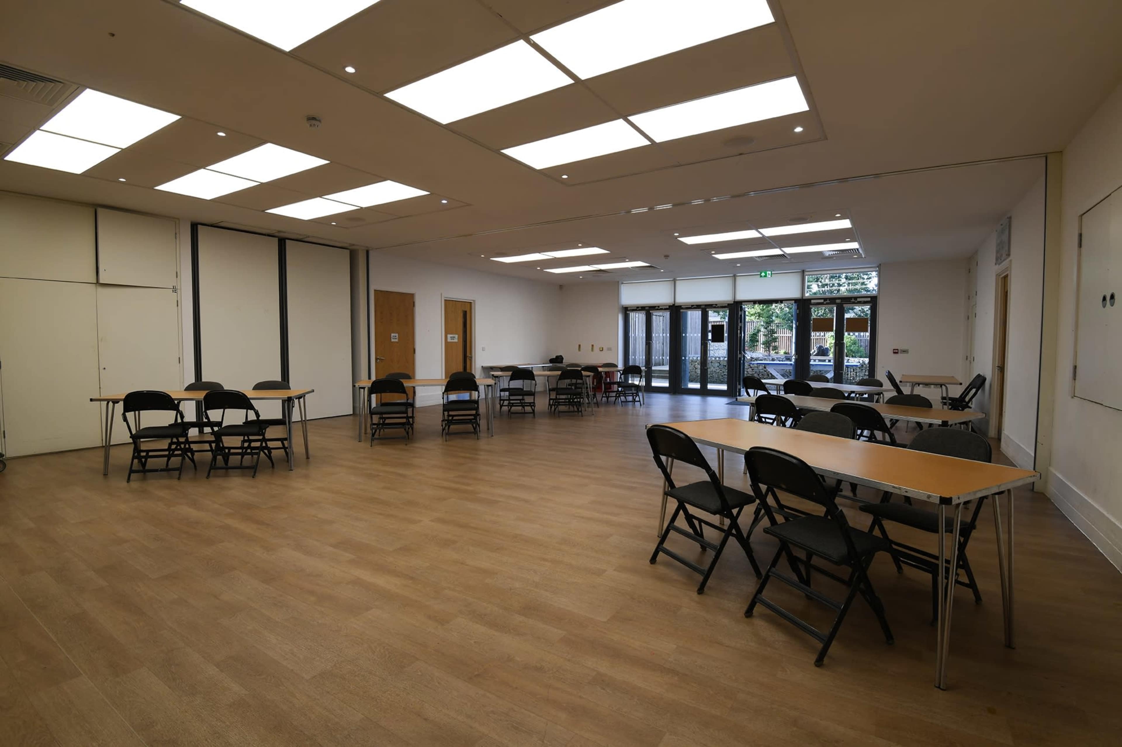 A spacious room with several folding tables and chairs arranged for seating, illuminated by overhead lights and featuring large windows that open to an outdoor area.