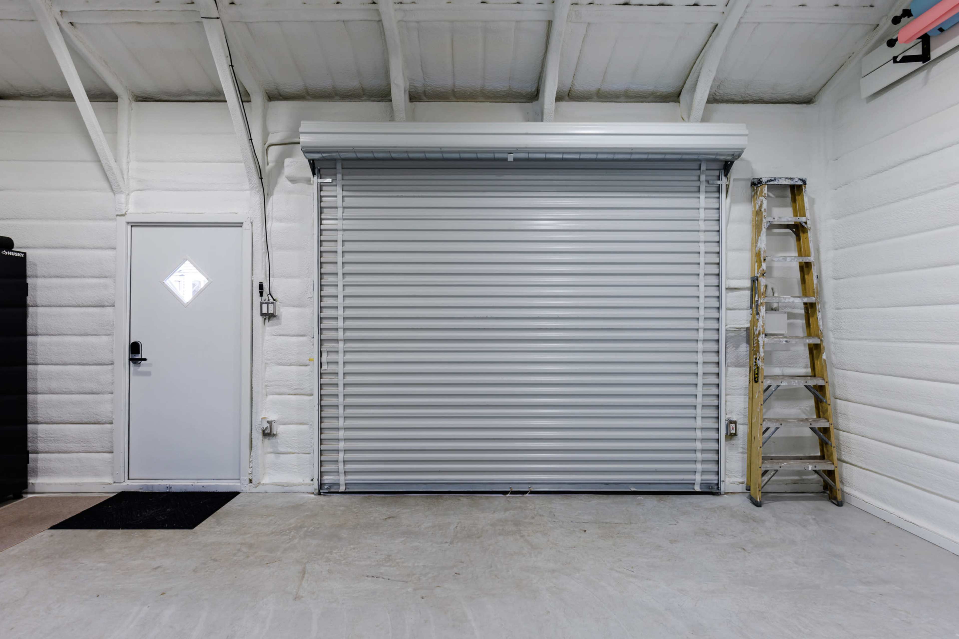 A garage interior with a closed rolling shutter door, a ladder against the wall, and a door on the left side.
