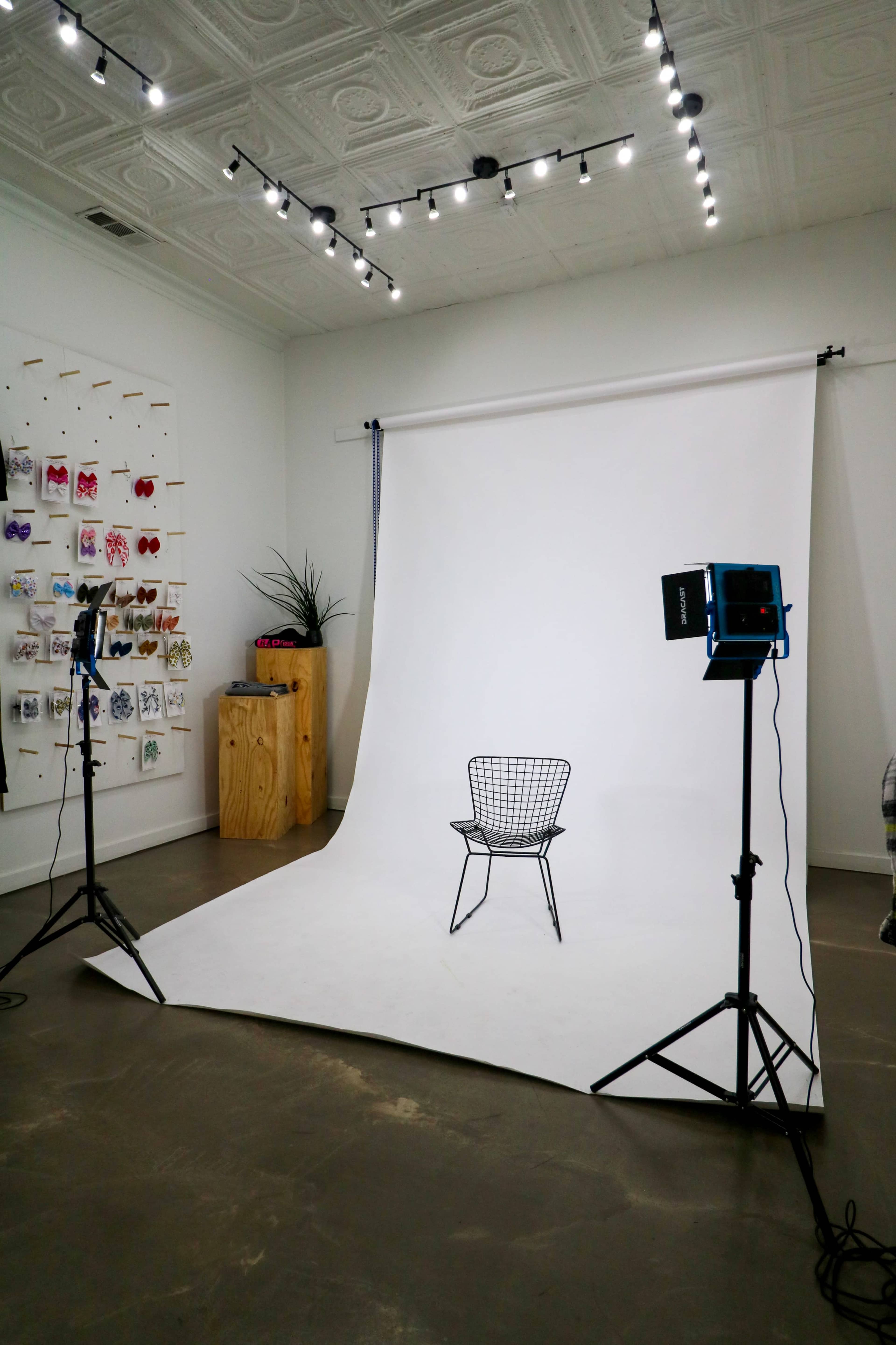 The image shows a minimalist photography studio setup with a white backdrop, a wire chair in the center, and two lighting stands on either side.