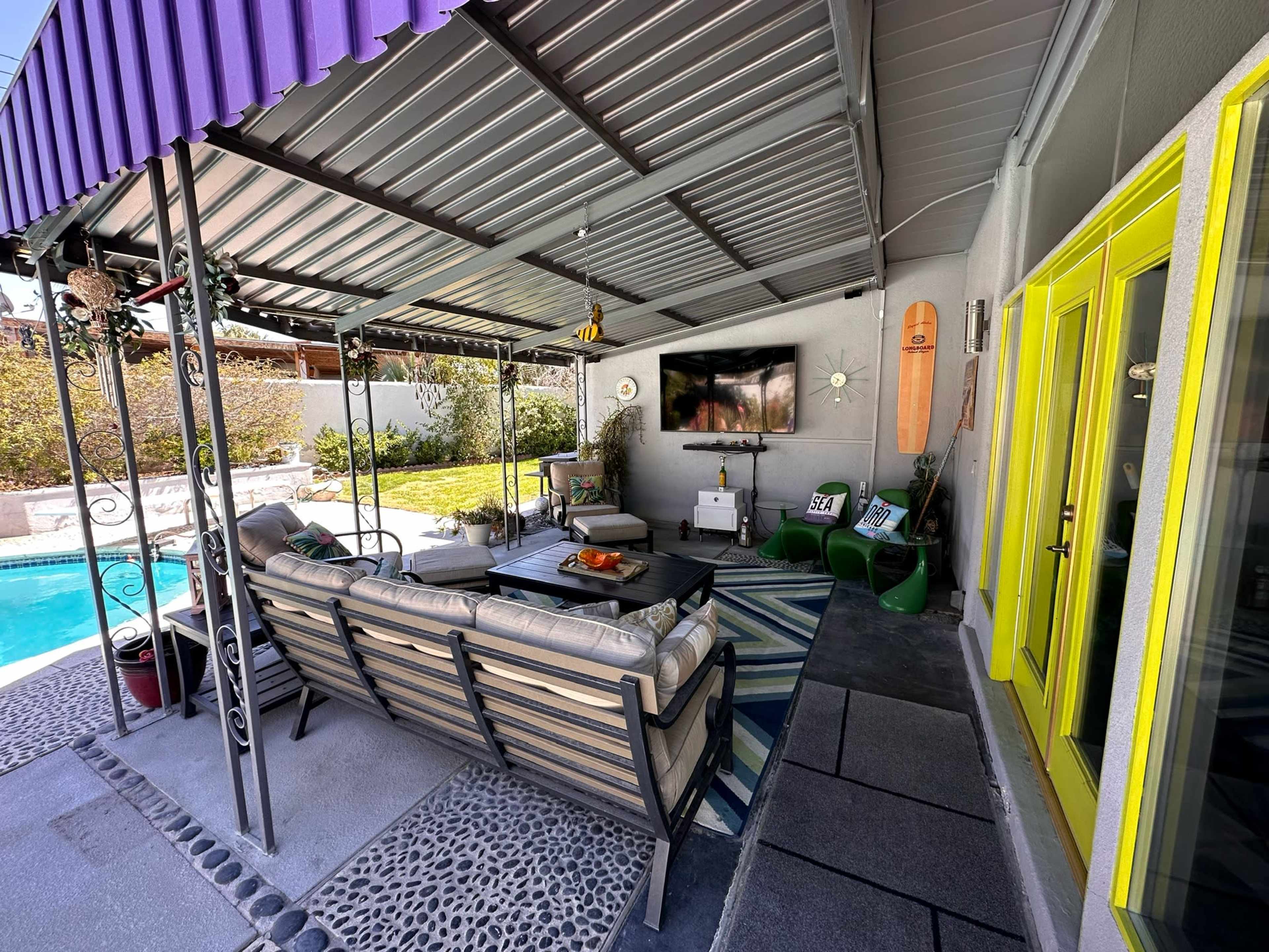 The image shows a covered outdoor patio with a seating area, a television mounted on the wall, and a view of a pool and lawn through large glass doors.