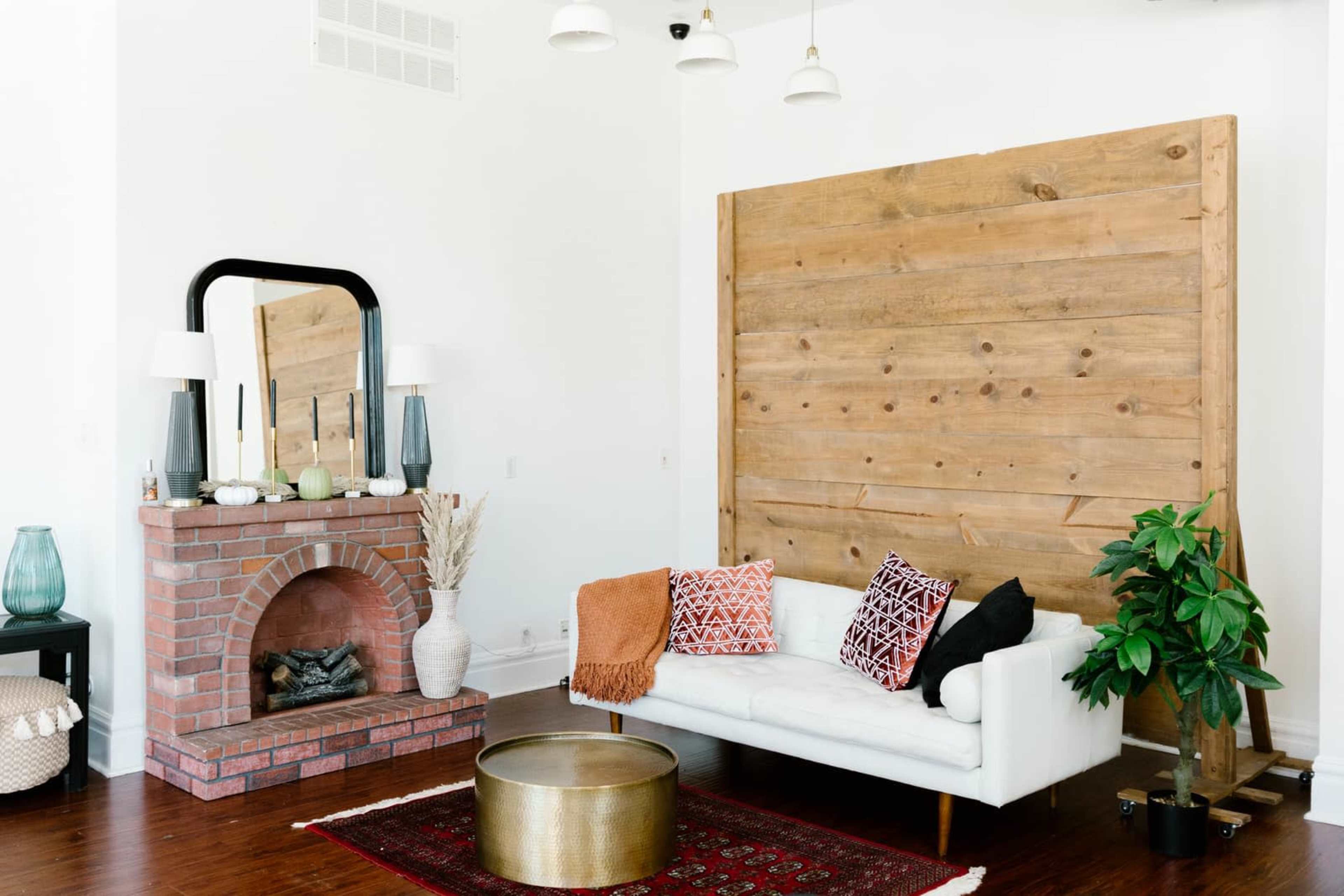 The living room features a white sofa with patterned pillows, a brick fireplace beneath a mirror, a wooden accent wall, and a potted plant beside a round coffee table.