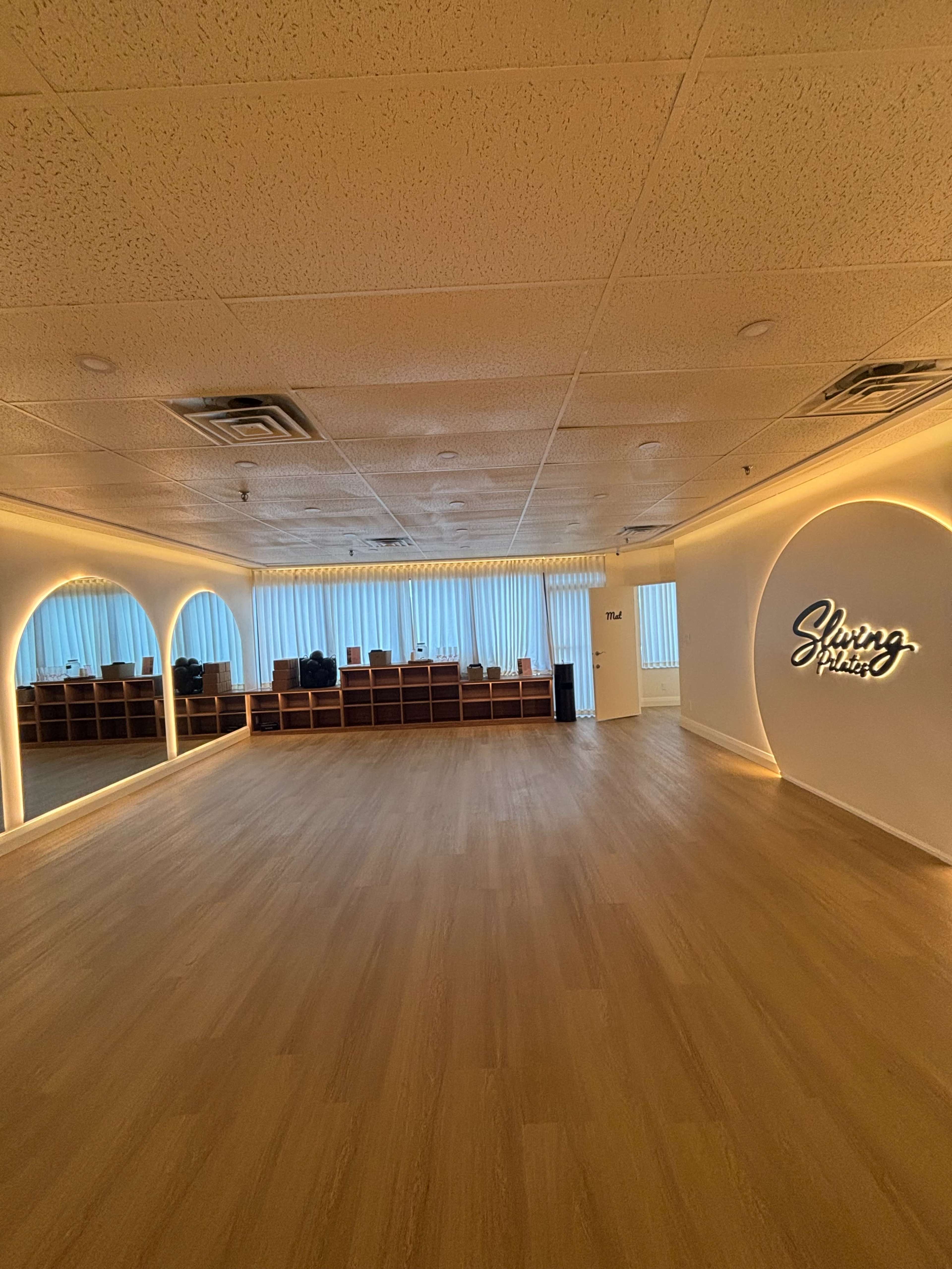 The image shows a spacious, well-lit room with wooden flooring, featuring a reception area with seating behind a counter and large windows letting in natural light.