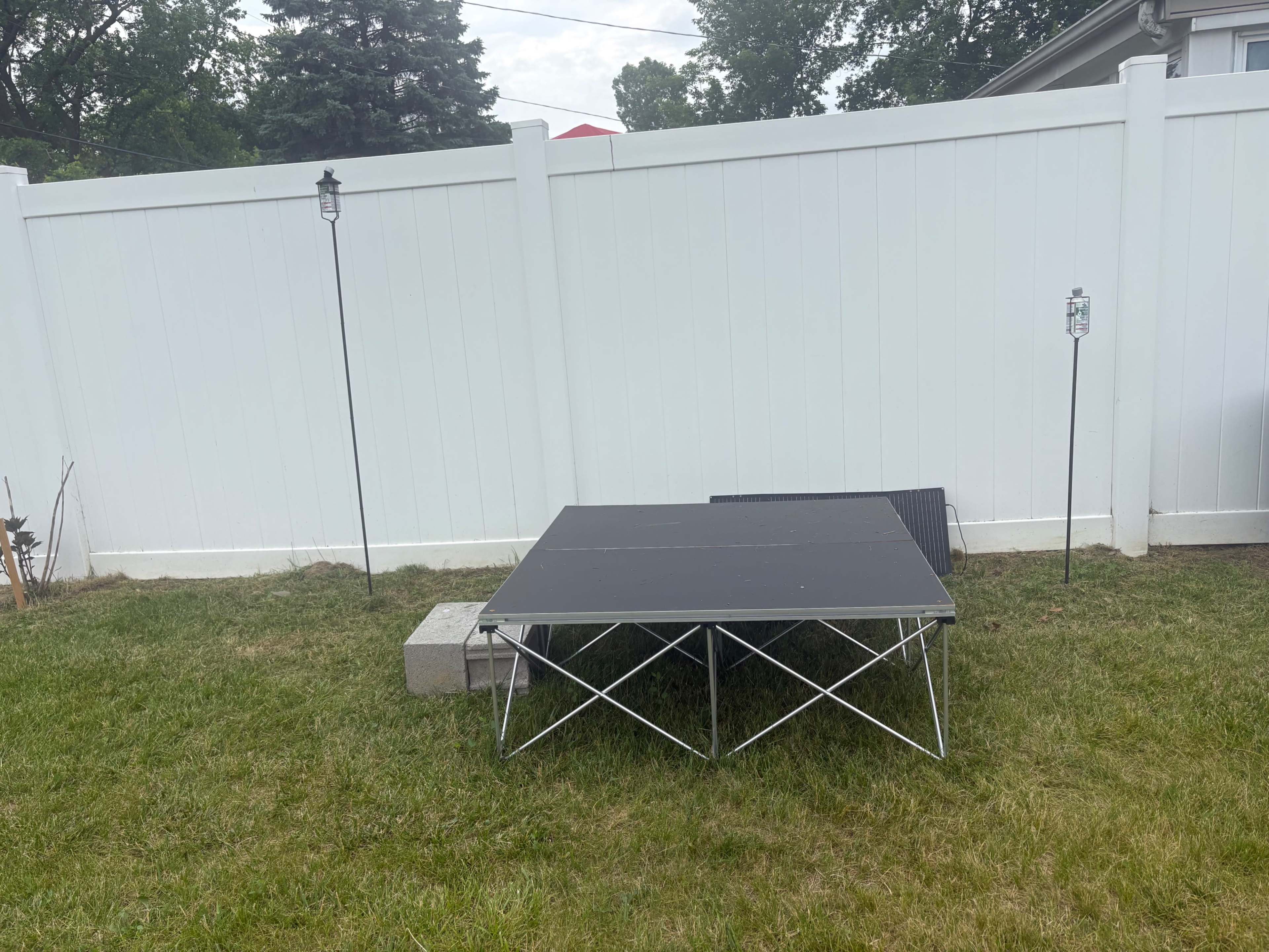 A black table is set up on grass beside a white fence, with two lanterns positioned on either side.