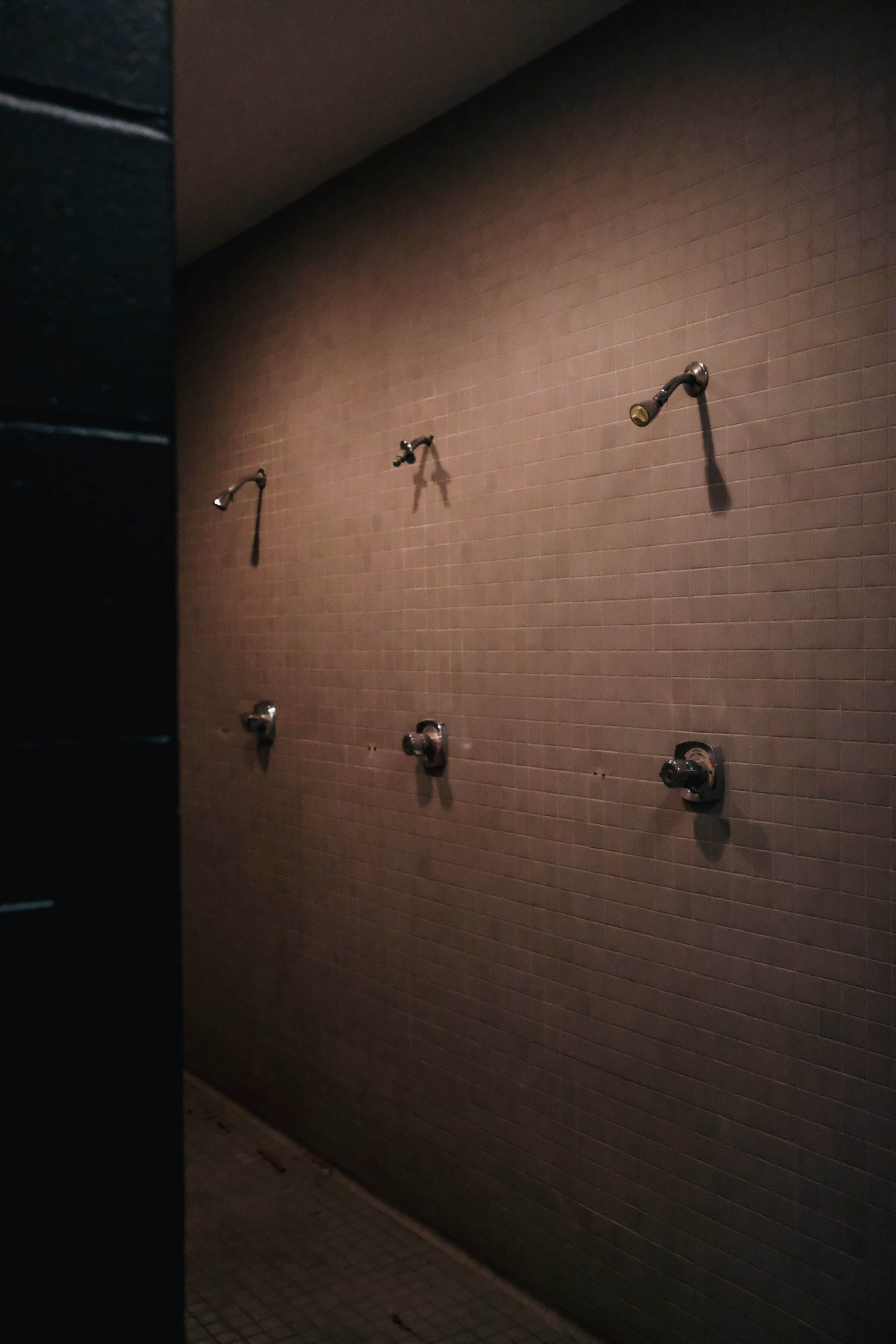 The image shows a dimly lit communal shower area with three showerheads mounted on a tiled wall.