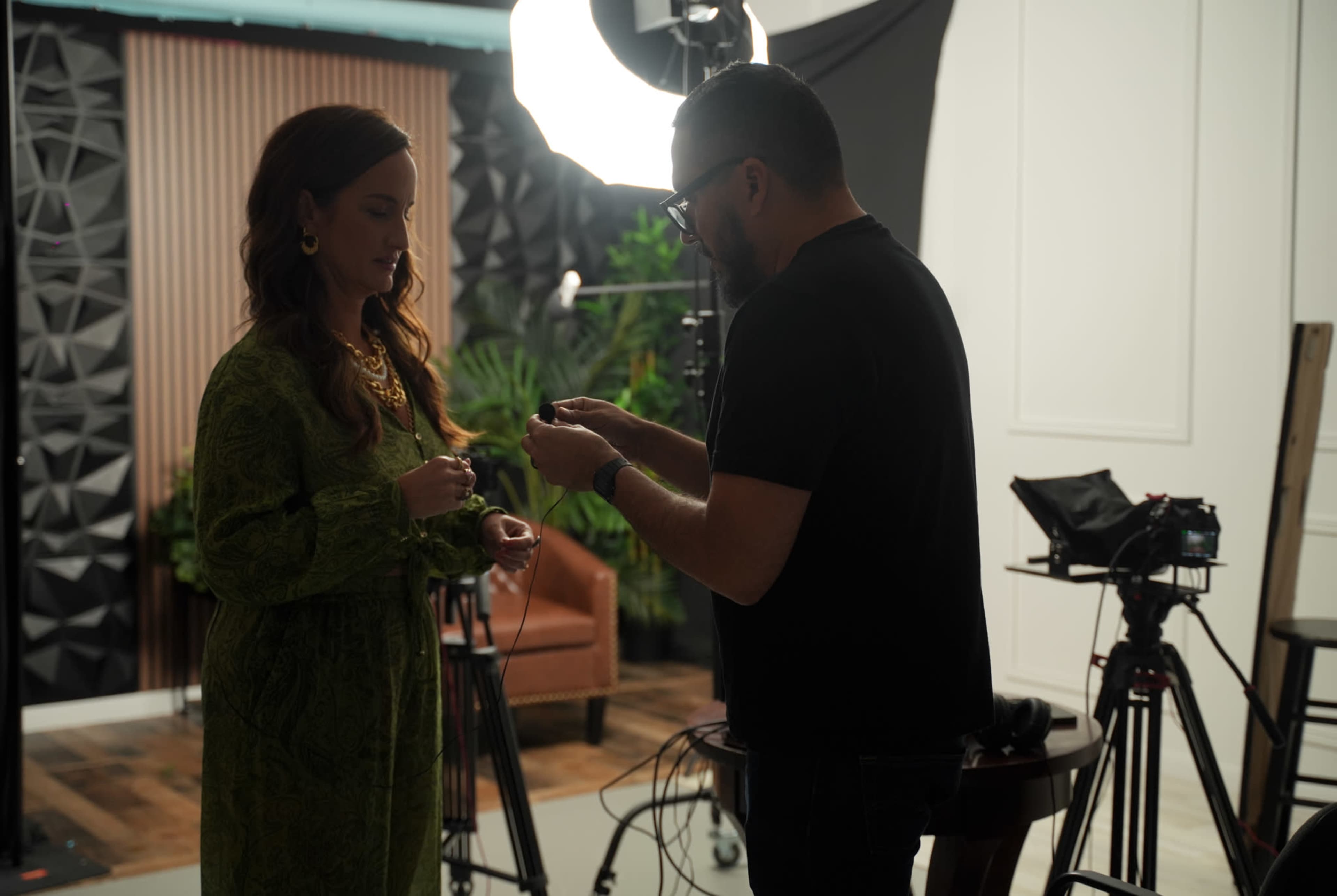 A woman in a green dress stands while a man adjusts something in her hands in a studio setting with lighting equipment and cameras.