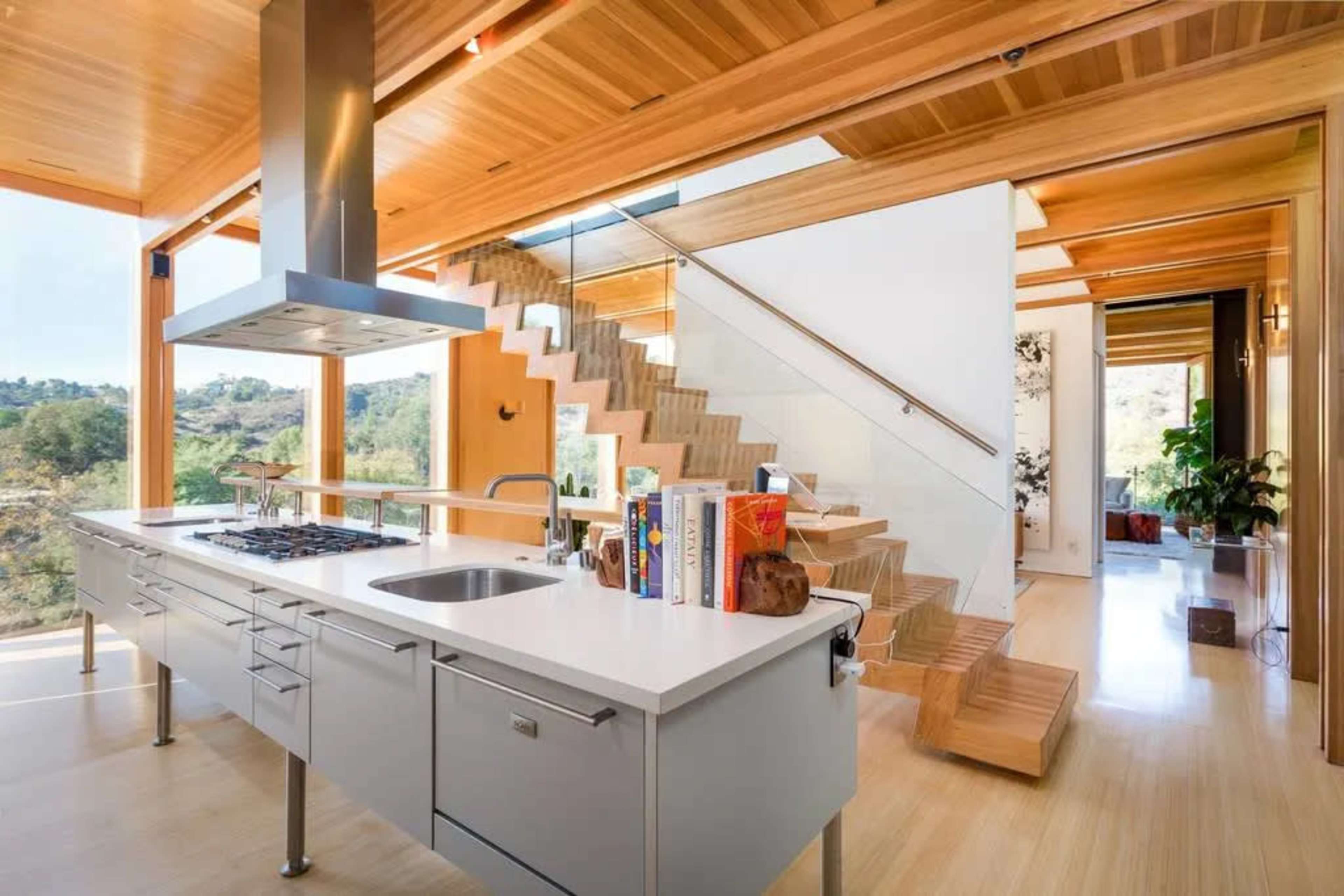 The image shows a modern kitchen with a central island and a staircase made of wood and glass leading to an upper level.