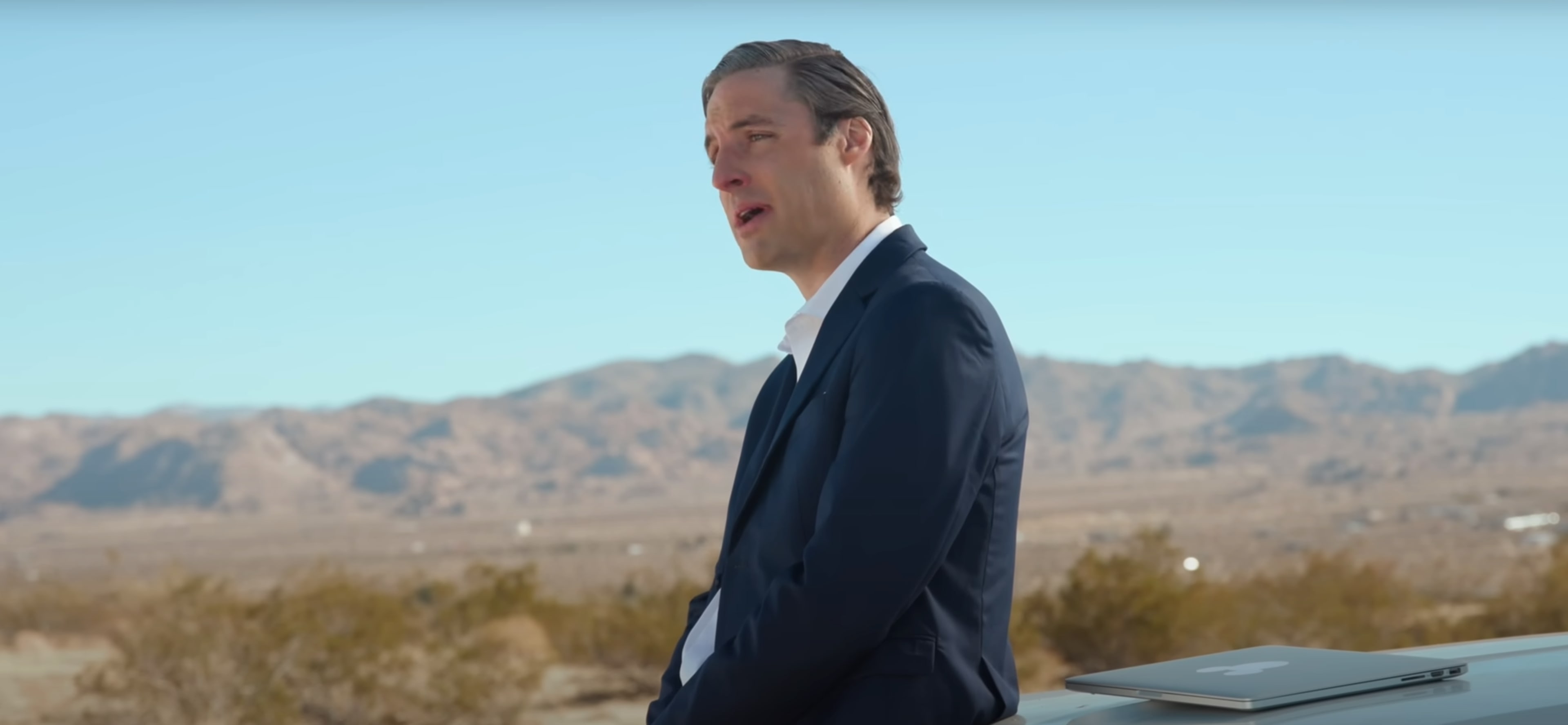 A man in a suit stands next to a laptop in a desert landscape with mountains in the background.