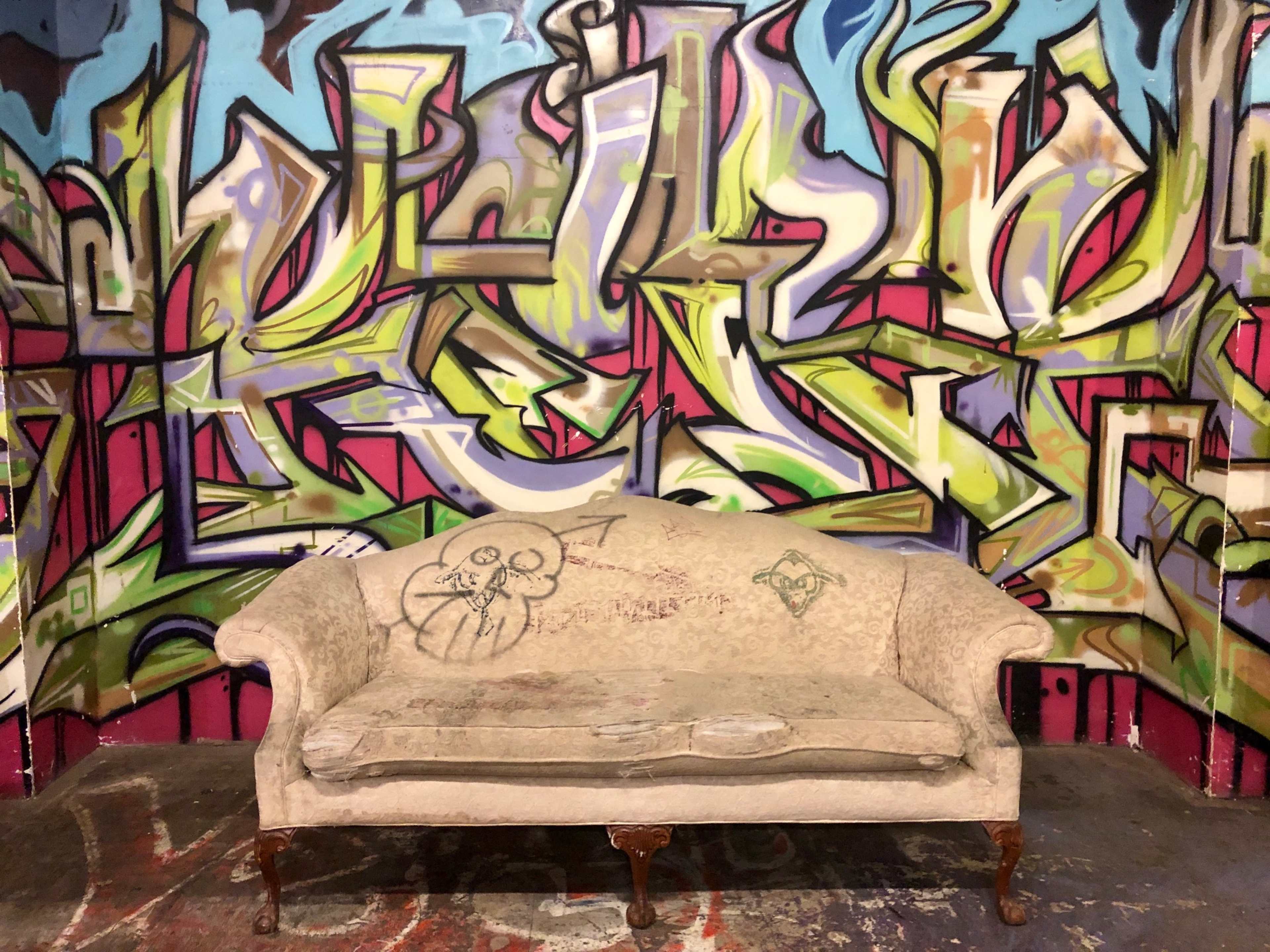A worn, light-colored couch sits against a vibrant, graffiti-covered wall.