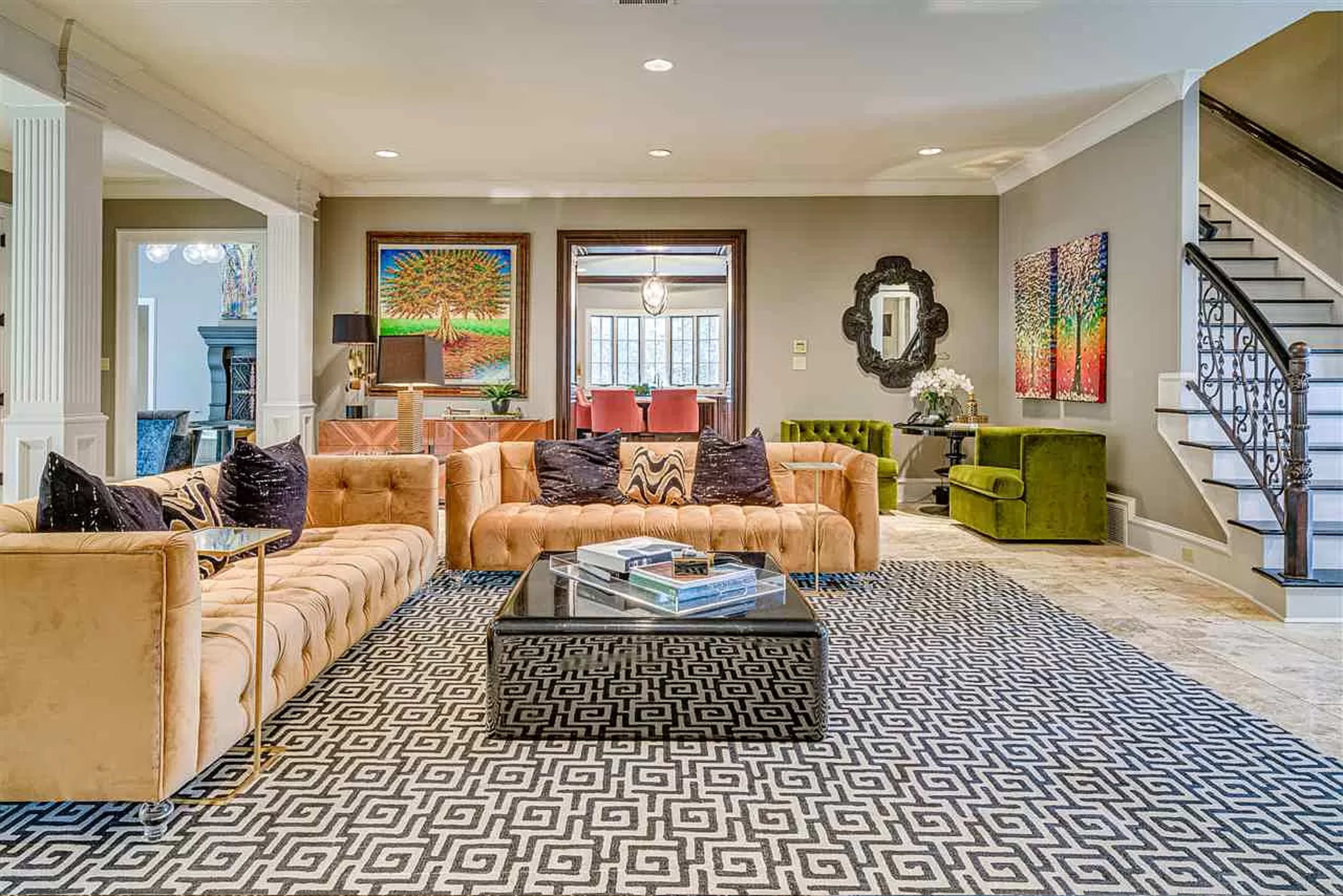 The living room features a large, beige tufted sofa, colorful wall art, a dark coffee table, and a patterned area rug, with a staircase visible in the background.