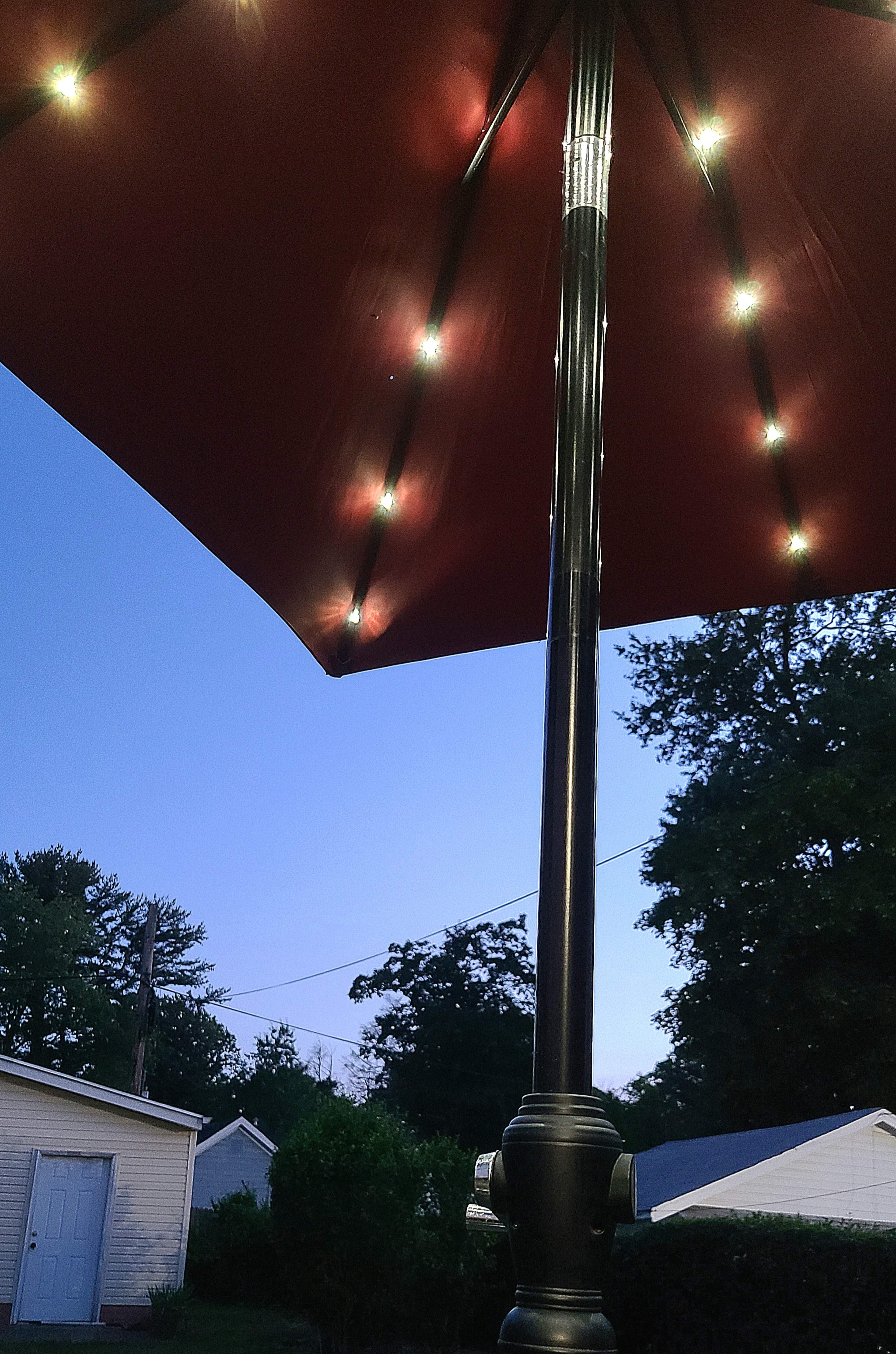 A lit patio umbrella stands over a backyard as dusk falls, with a house and trees in the background.