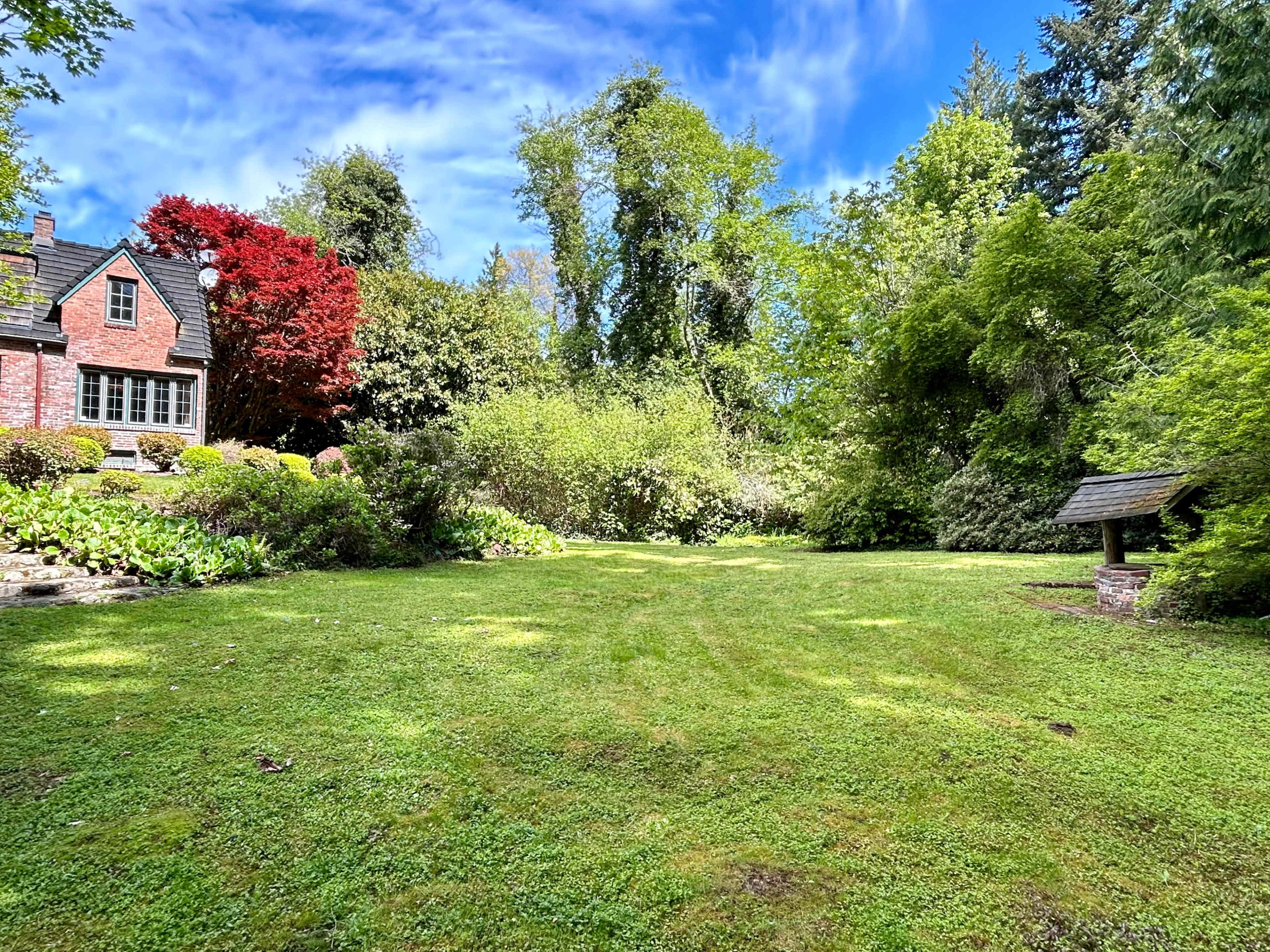 A landscaped yard with lush green grass, surrounded by trees and shrubs, and featuring a brick house and a small well structure.