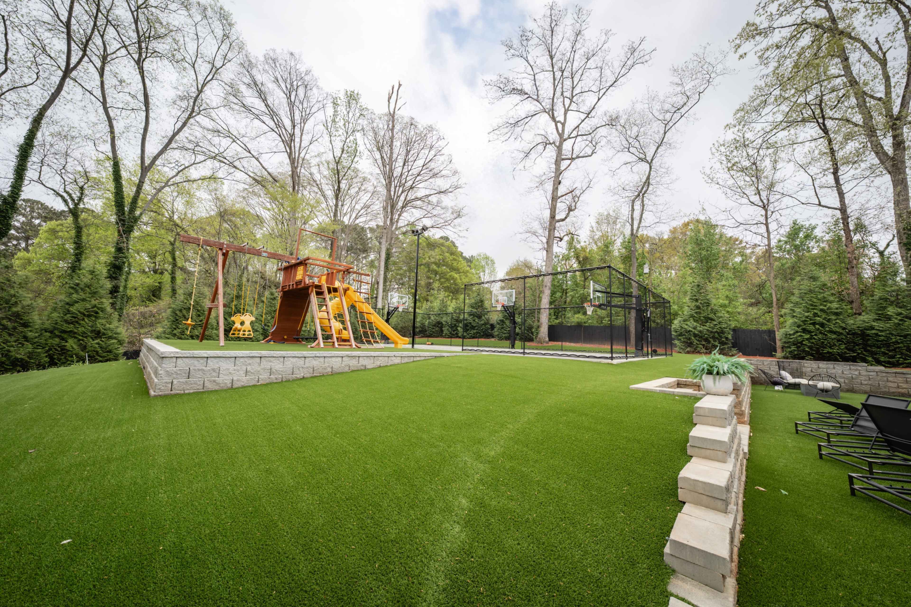 The image shows a backyard featuring a wooden playset with a slide and swings, an enclosed sports court, and well-maintained artificial grass surrounded by trees.