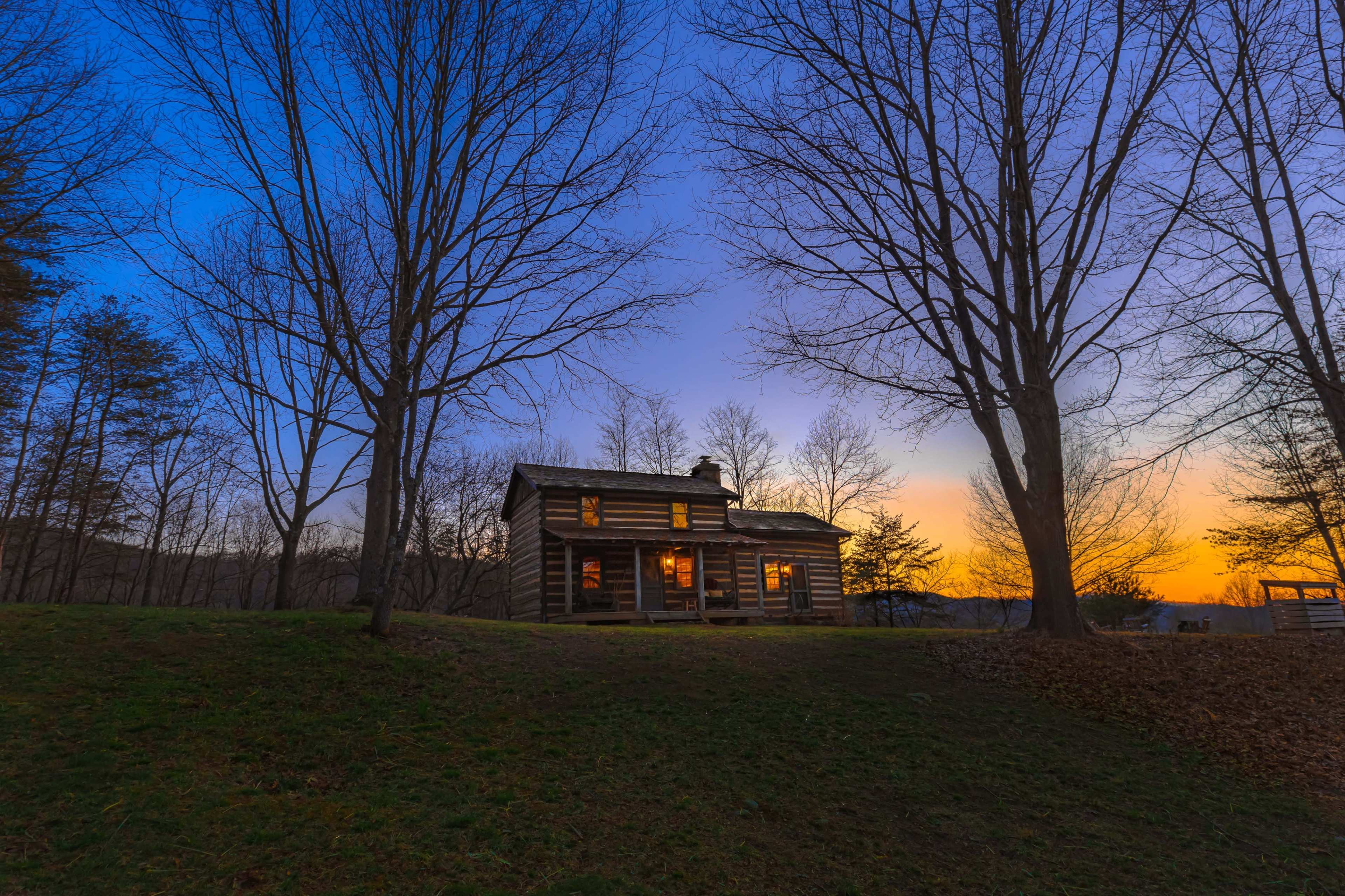 A log cabin with illuminated windows stands on a hillside at sunset, surrounded by bare trees.