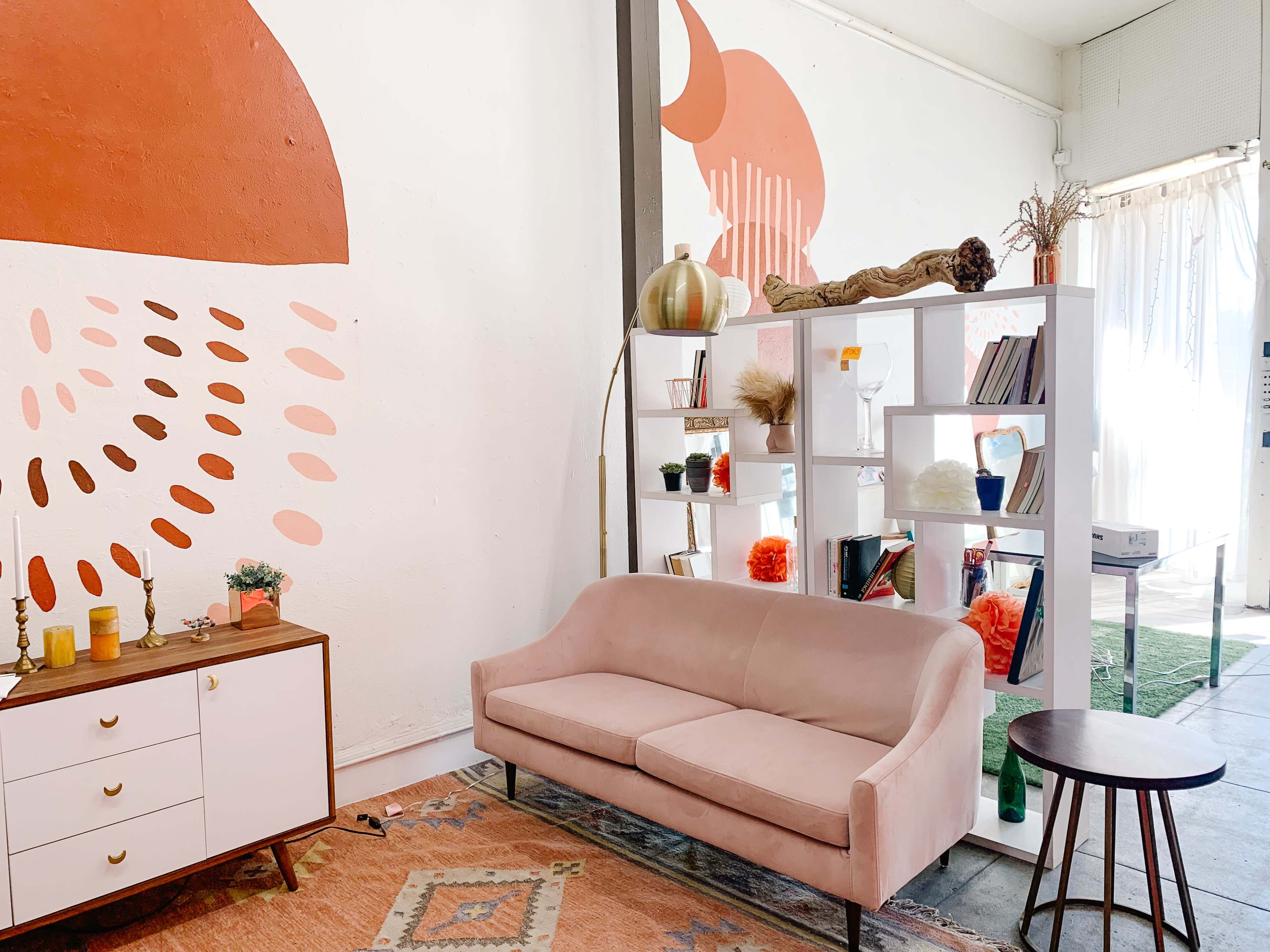 A modern interior featuring a pink sofa, a shelving unit with decorative items and books, and a wall painted with geometric patterns in warm tones.