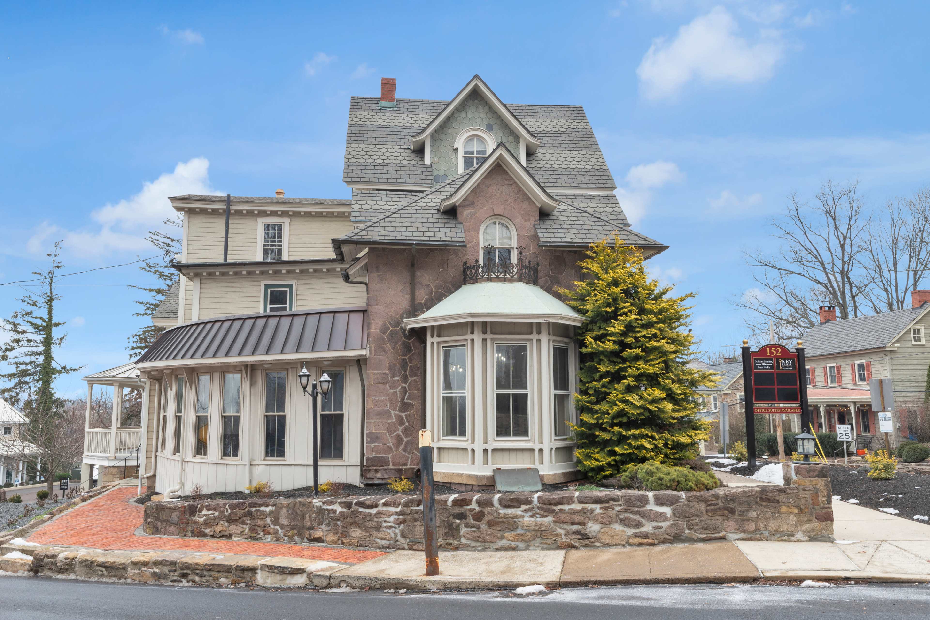 The image shows a historic two-story house with a gabled roof, a bay window, and a stone foundation, located at the corner of a street.