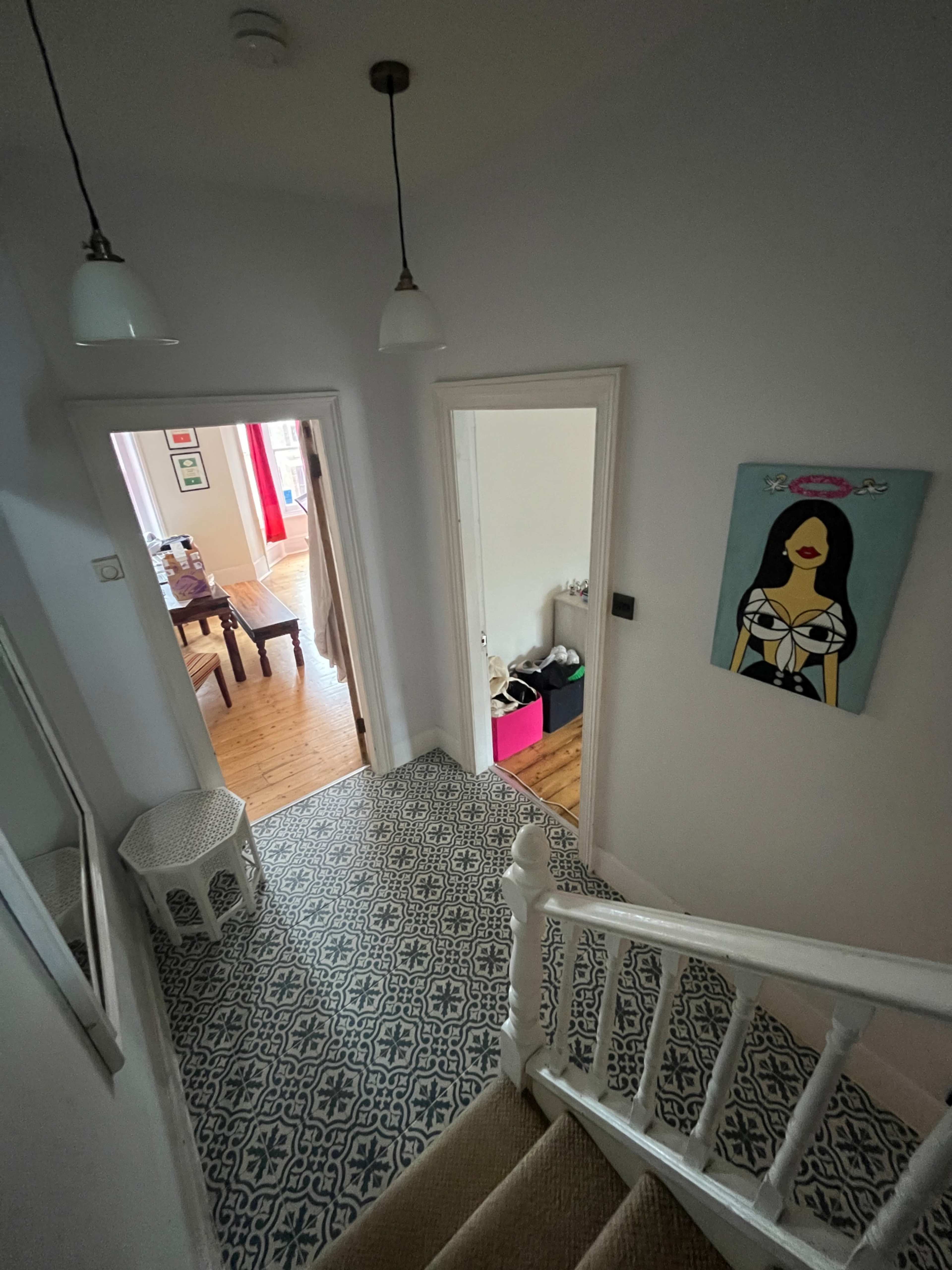 The image shows a staircase leading down to a hallway with a patterned floor, a doorway to a room, and a painting on the wall.