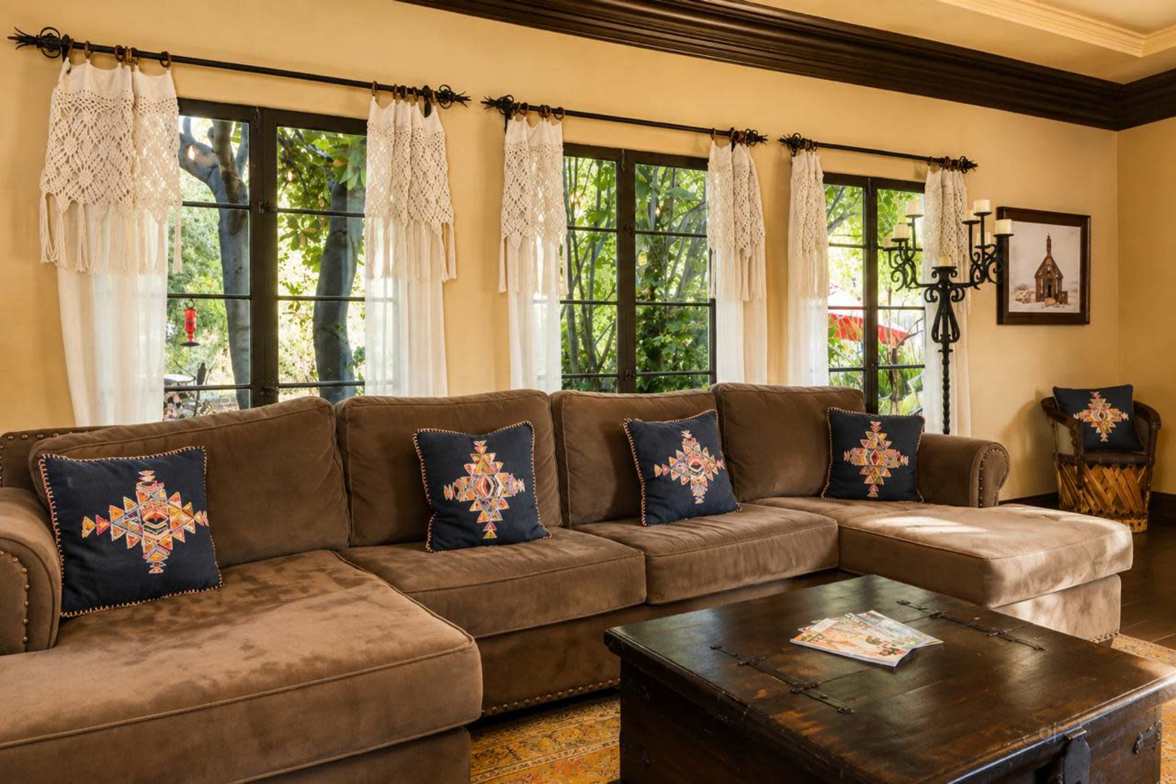 The image shows a cozy living room with a brown sectional sofa adorned with colorful pillows, large windows letting in natural light, and a dark wooden coffee table.
