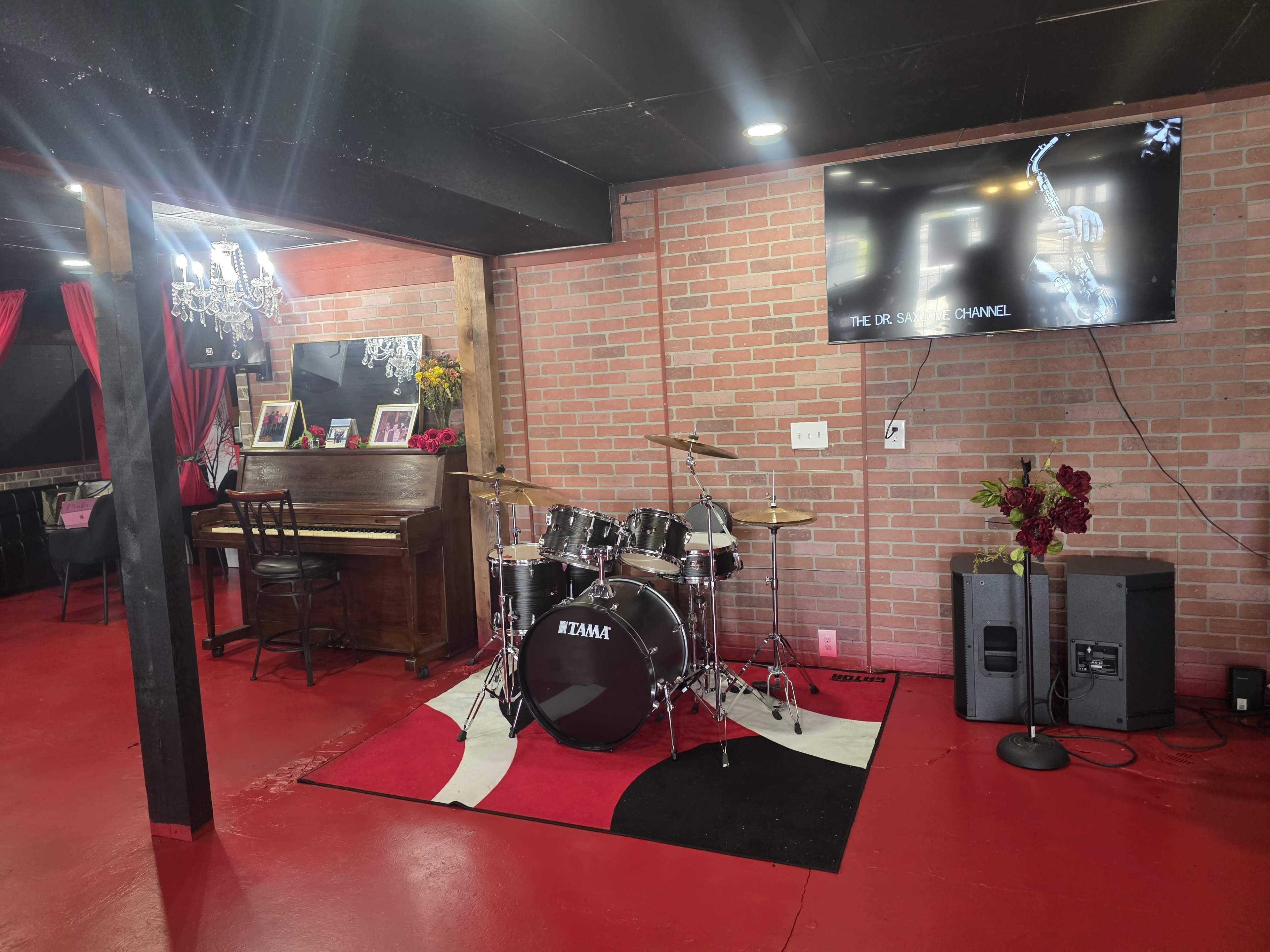 A music room features a drum set, a piano, a television mounted on a brick wall, and red flooring.