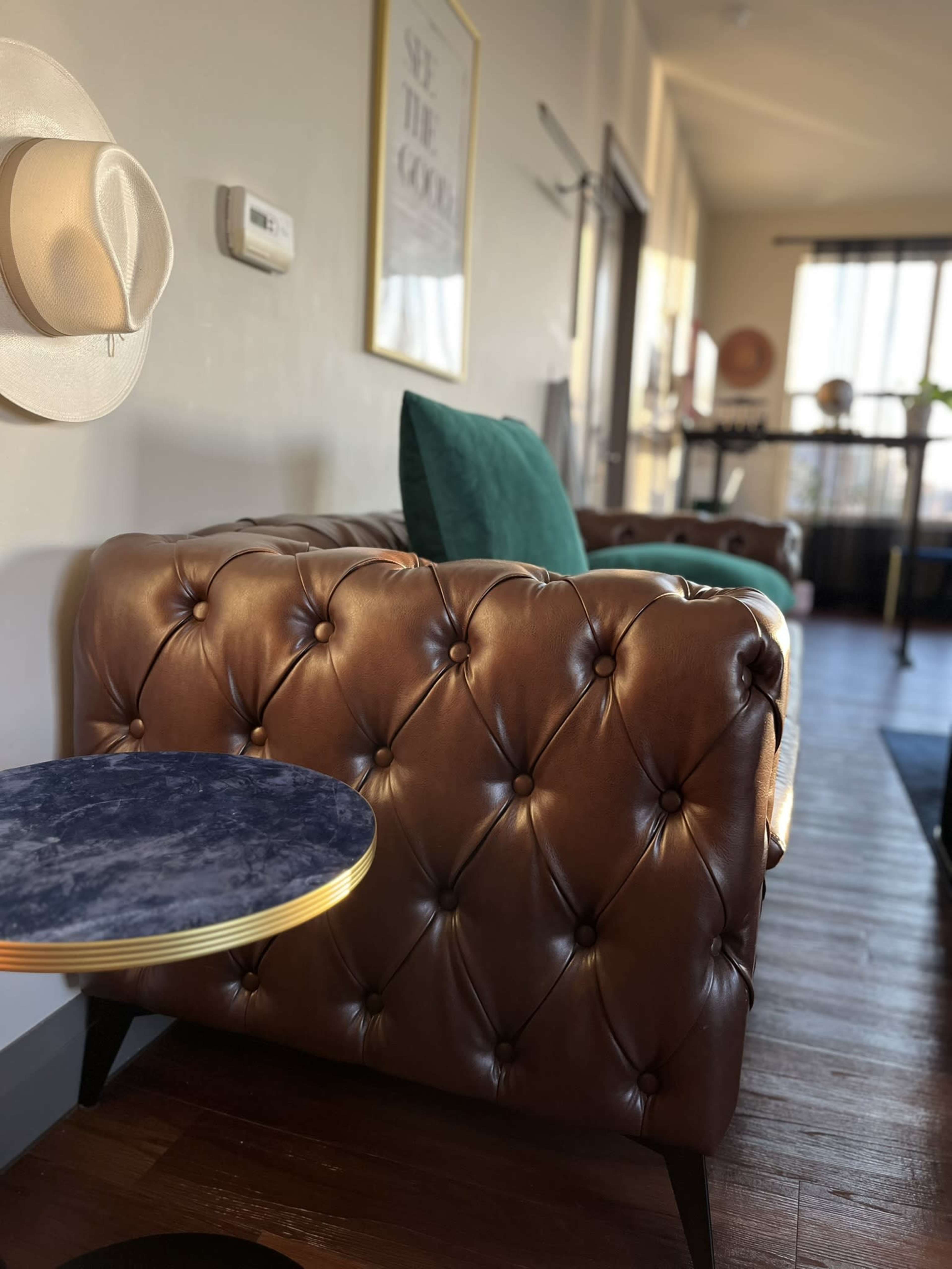The image features a brown tufted leather sofa, a green upholstered chair, and a small round table with a blue marble top in a well-lit interior space.