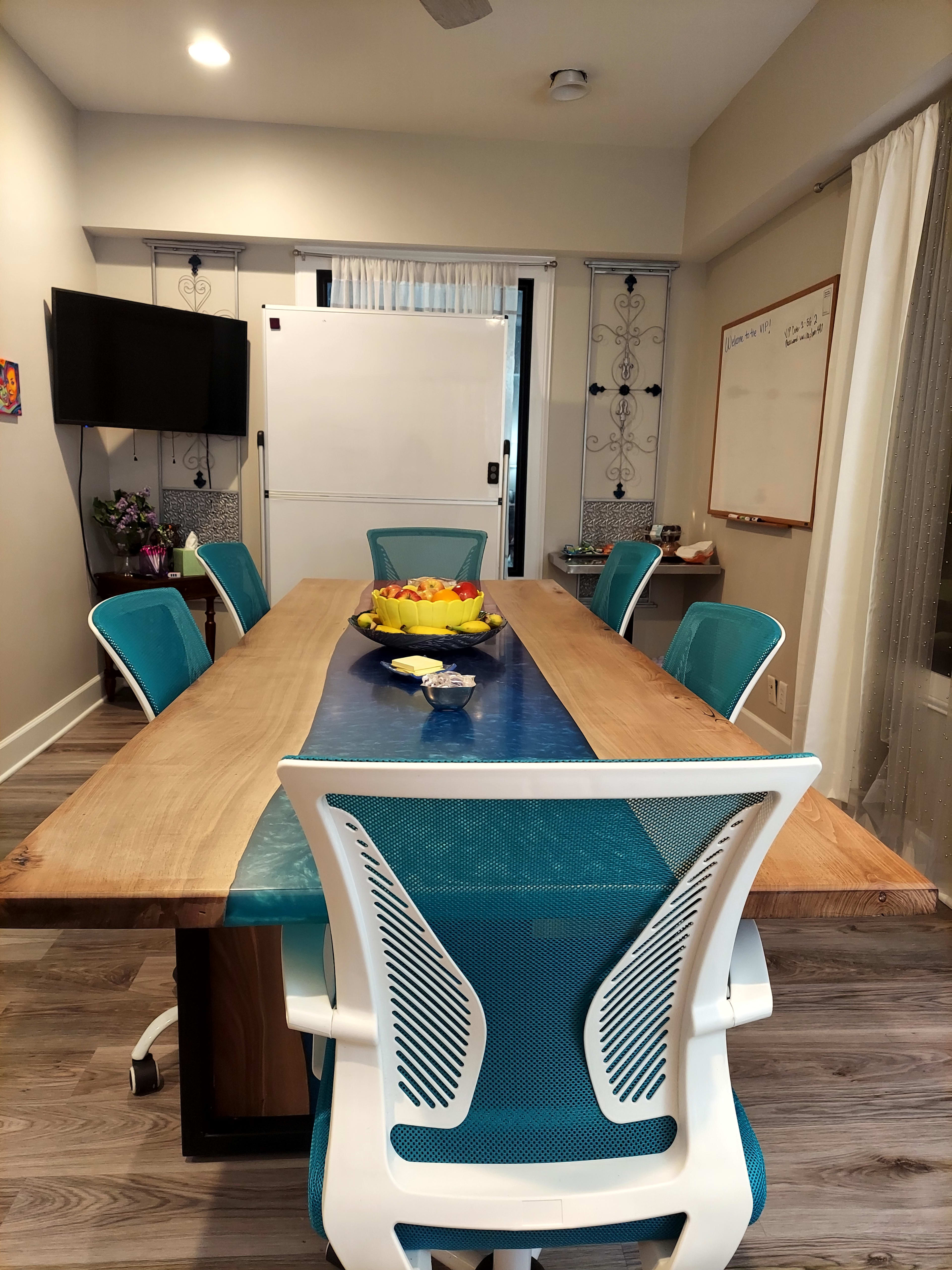 A conference room features a long wooden table with turquoise chairs, a fruit bowl at the center, and a television on one wall.