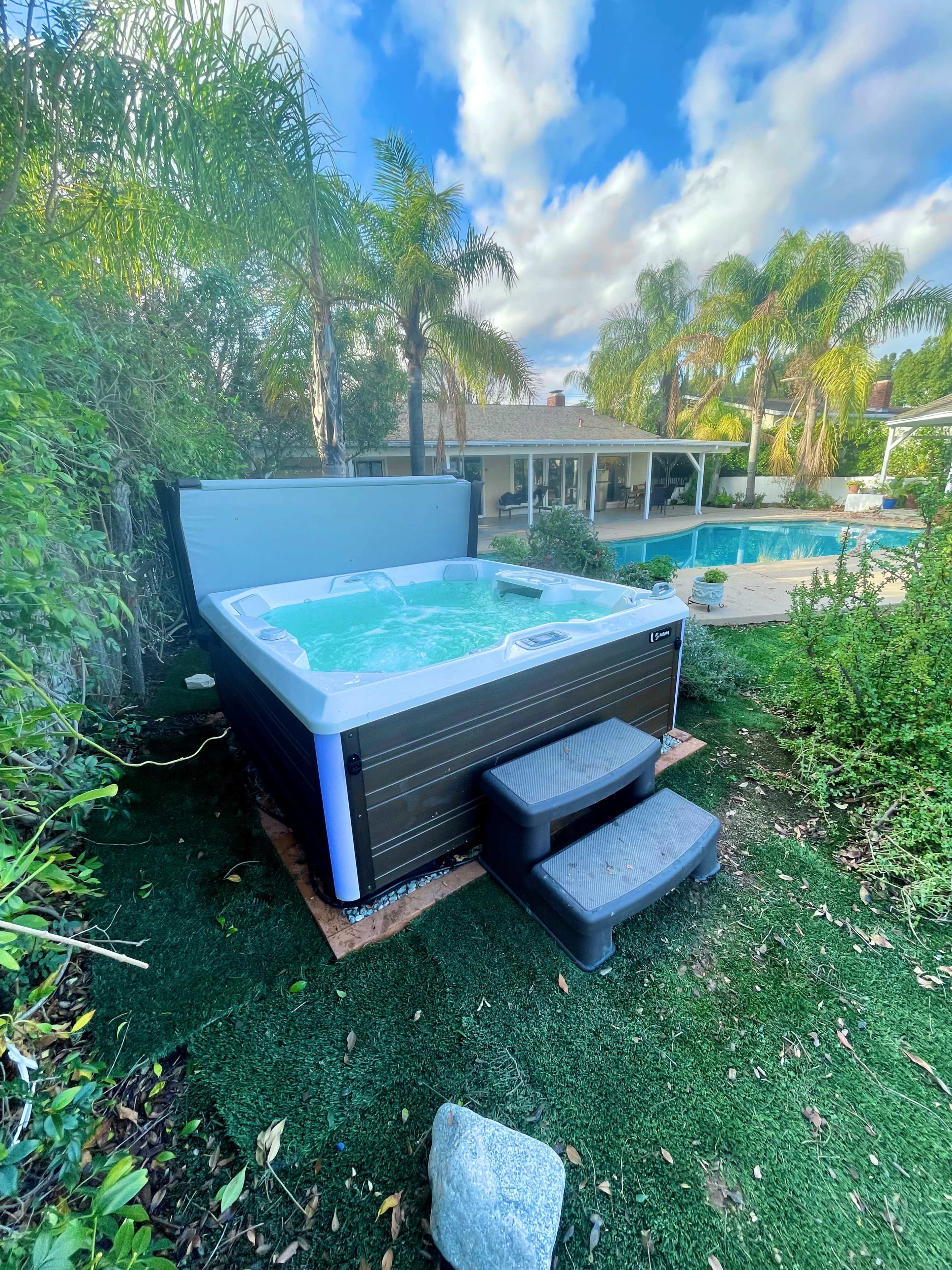 A hot tub is placed beside a swimming pool, surrounded by greenery and palm trees.