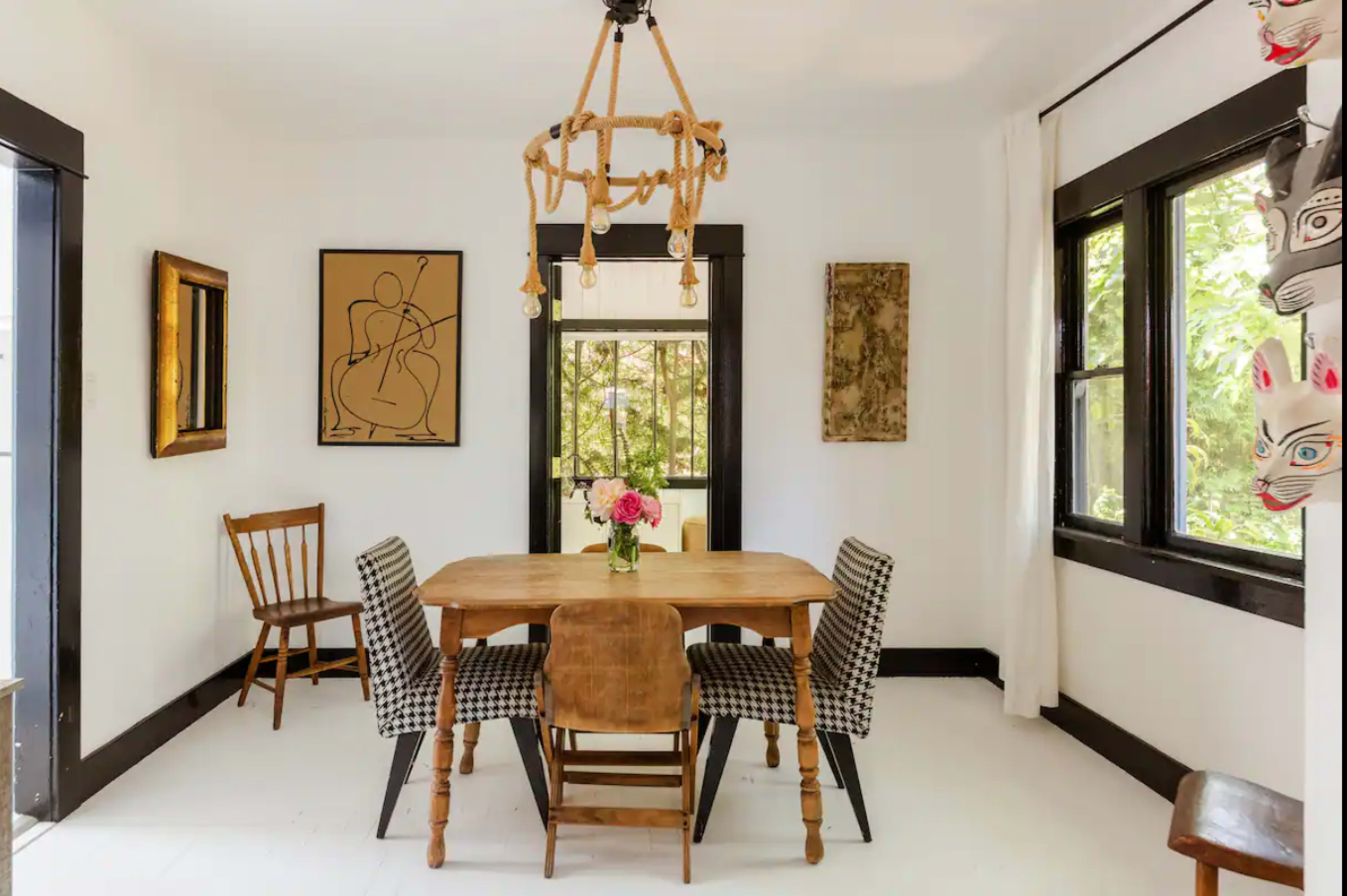 A wooden dining table is surrounded by chairs in a bright room with large windows and art on the walls.