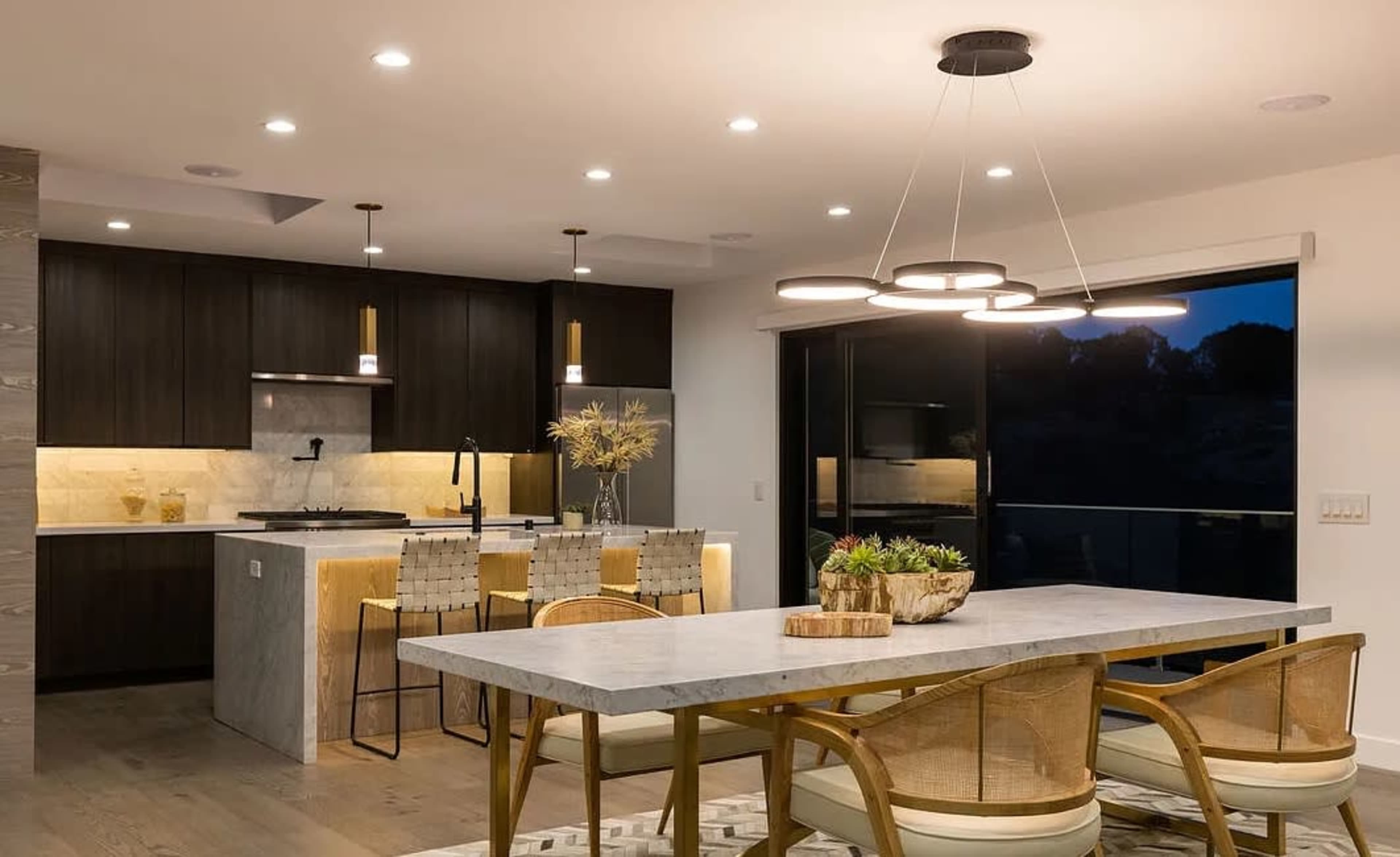 The image shows a modern kitchen and dining area featuring a marble dining table, woven chairs, and a sleek kitchen with dark cabinetry and pendant lighting.