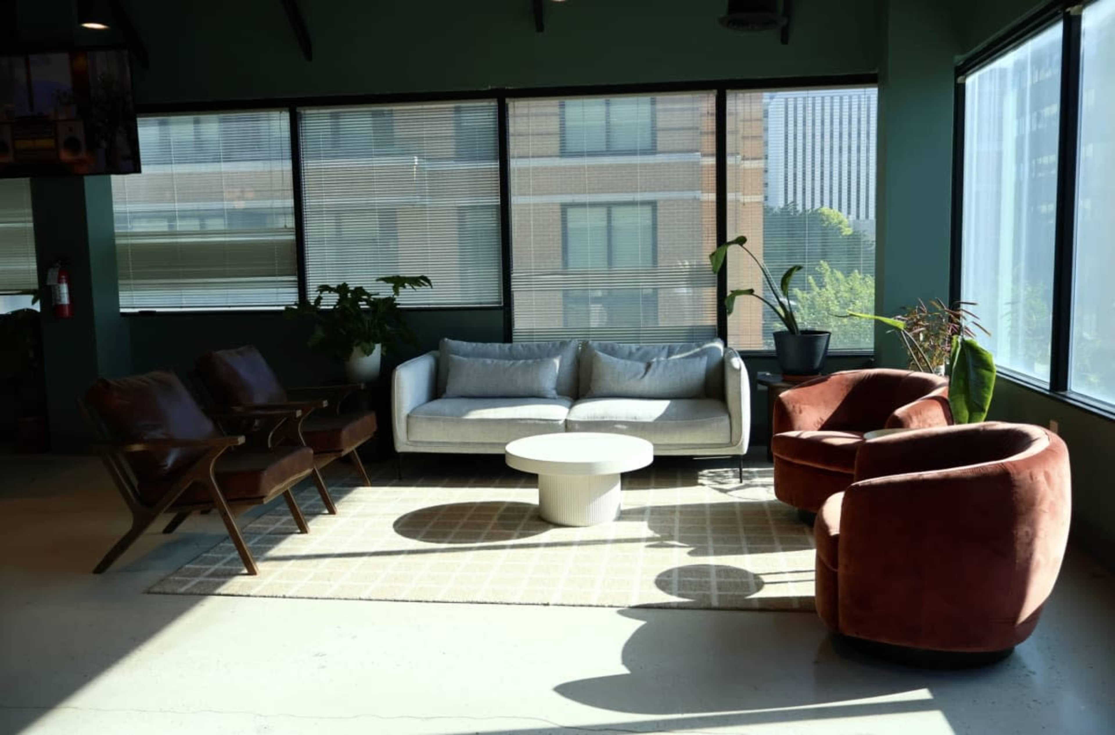 The image shows a bright room featuring a light gray couch, two red armchairs, a circular white coffee table, and various indoor plants near large windows.