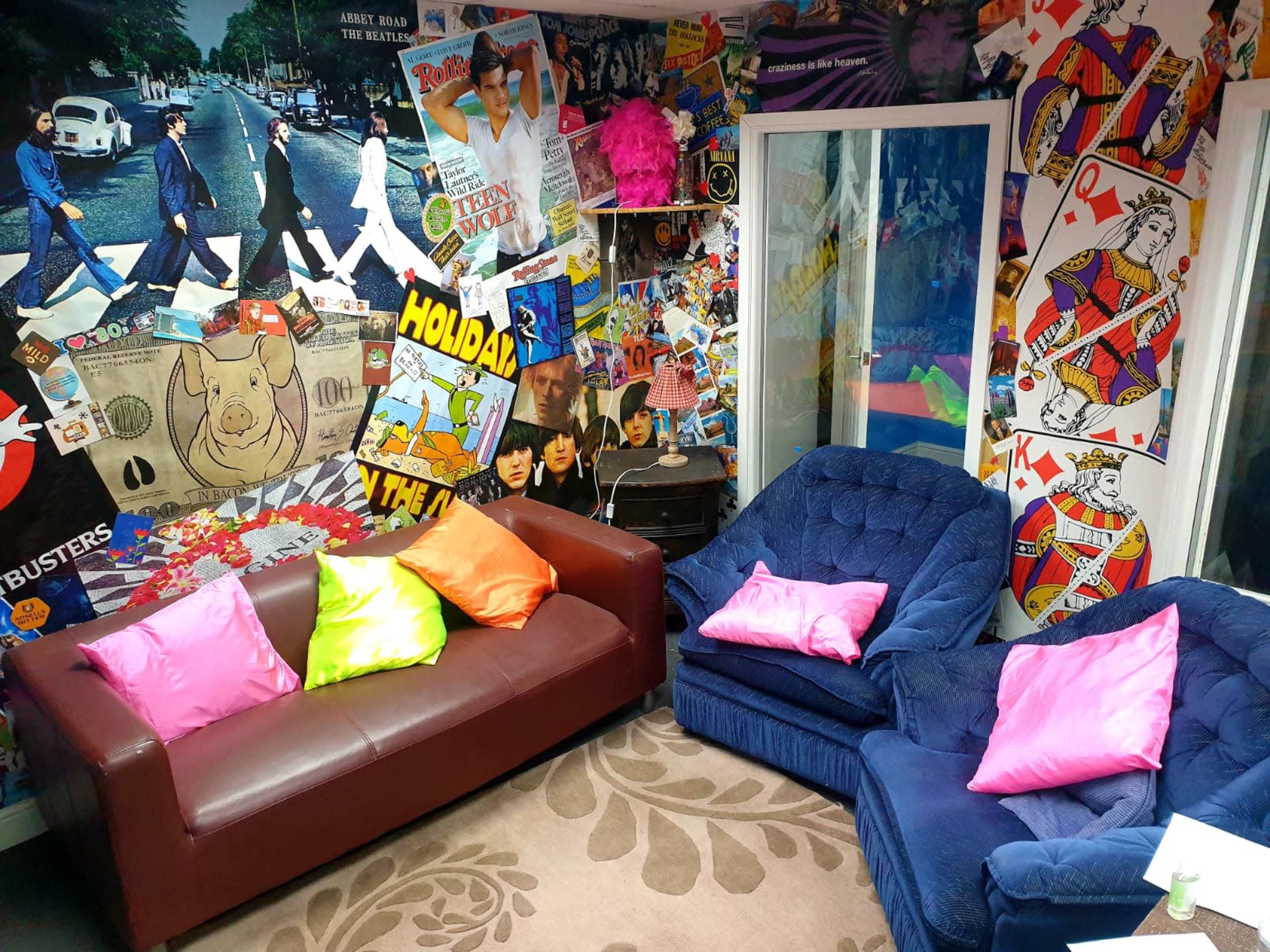 The room features two sofas, one red and one blue, surrounded by a colorful collage of posters and memorabilia on the walls.
