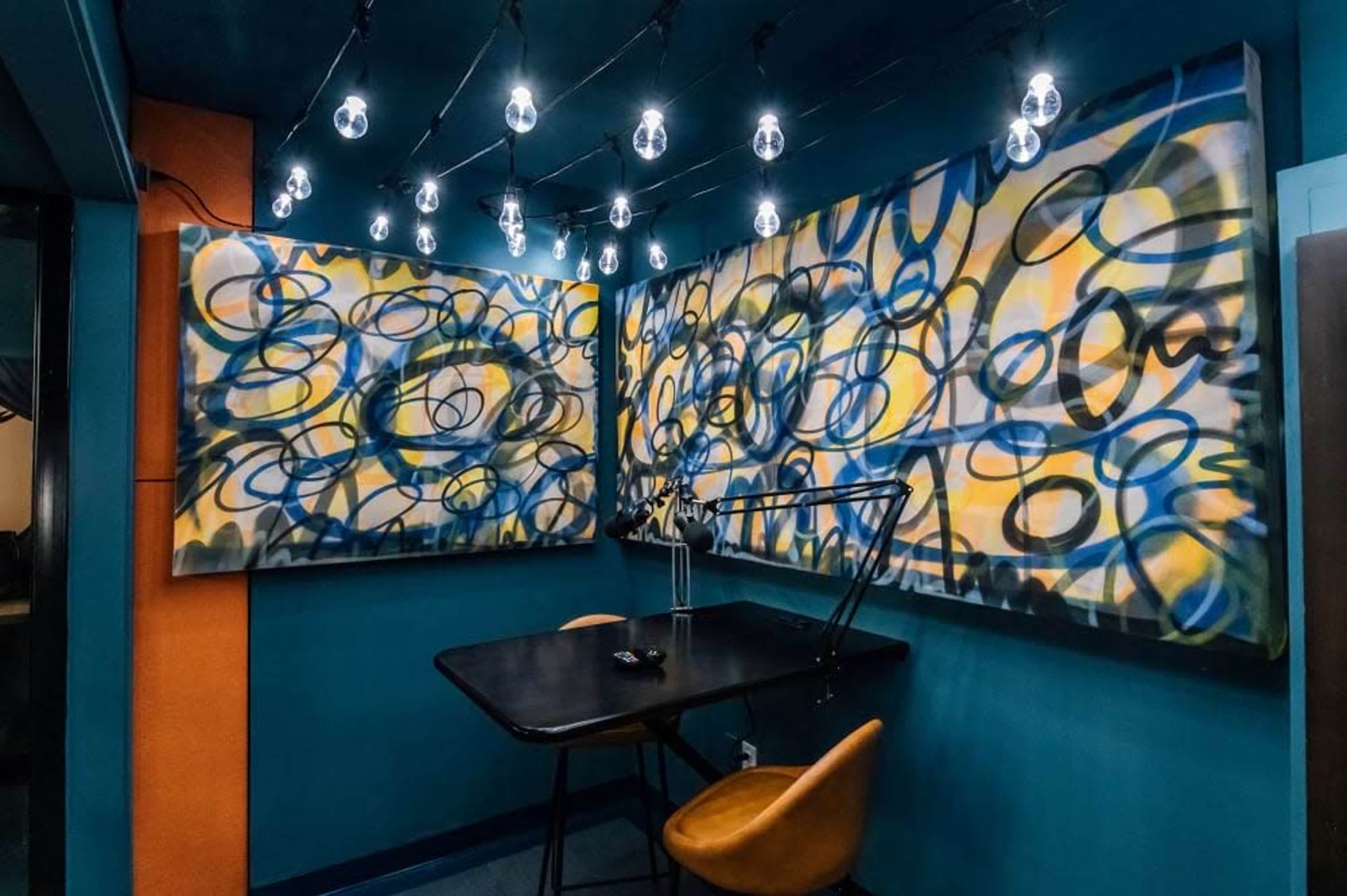 The image shows a small room with dark blue walls, abstract art on two walls, a black table with a chair, and bright overhead lights.