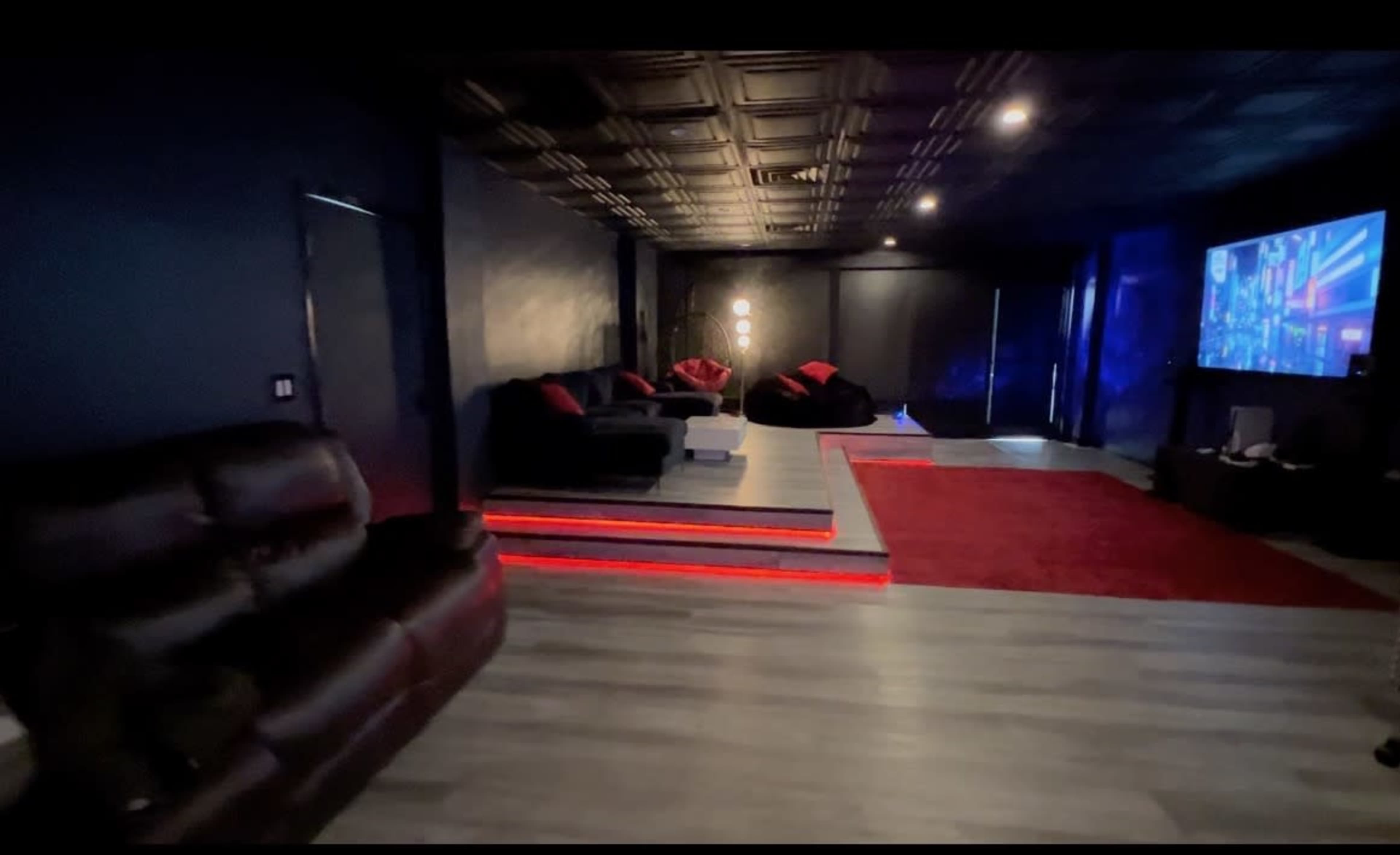 A dimly lit home theater features a raised seating area with dark couches and red accents, alongside a large screen on one wall.