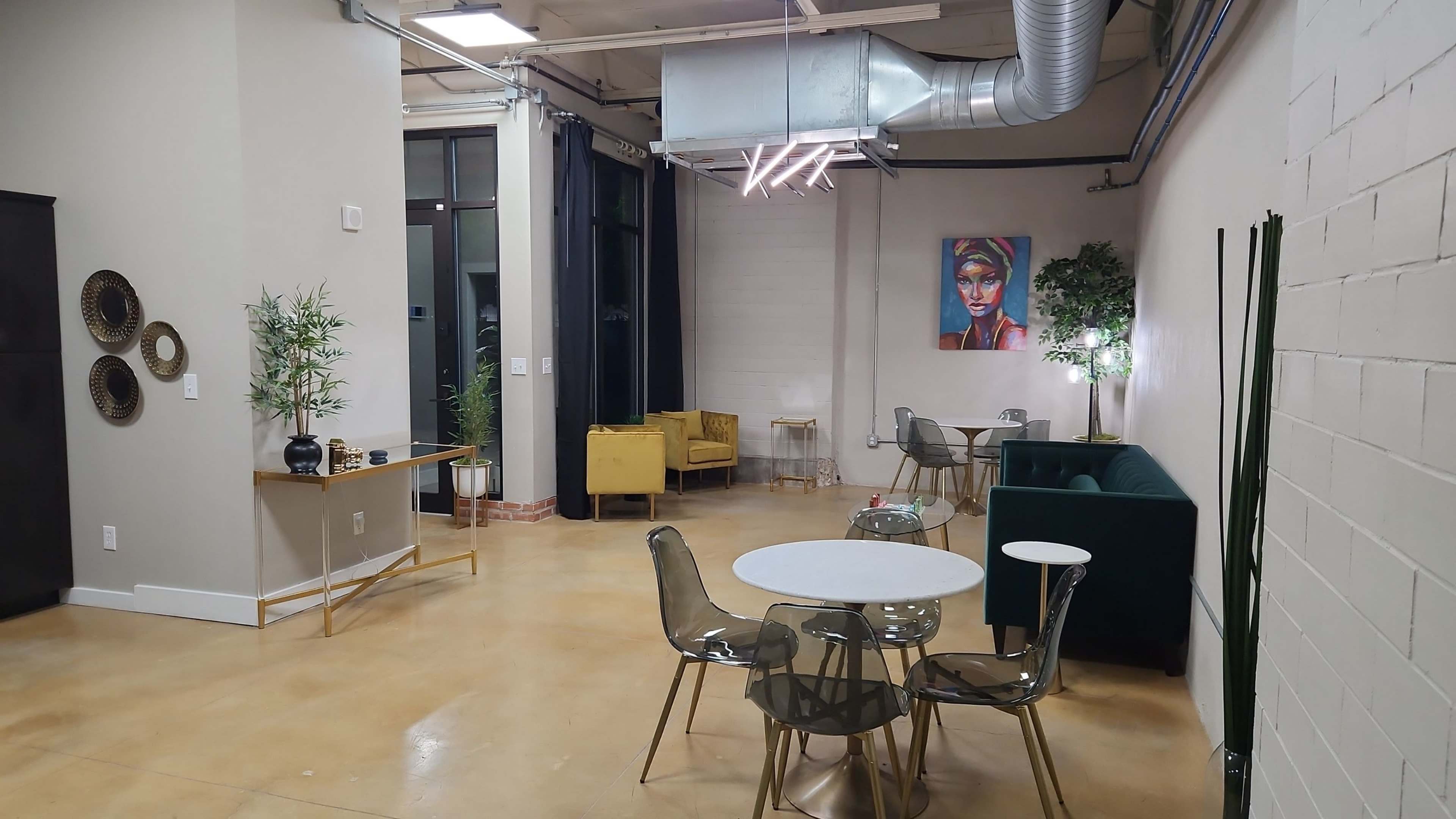 A modern, open interior space with a seating area that includes a green sofa, yellow chair, and several round tables surrounded by transparent chairs, along with art on the wall and plants.