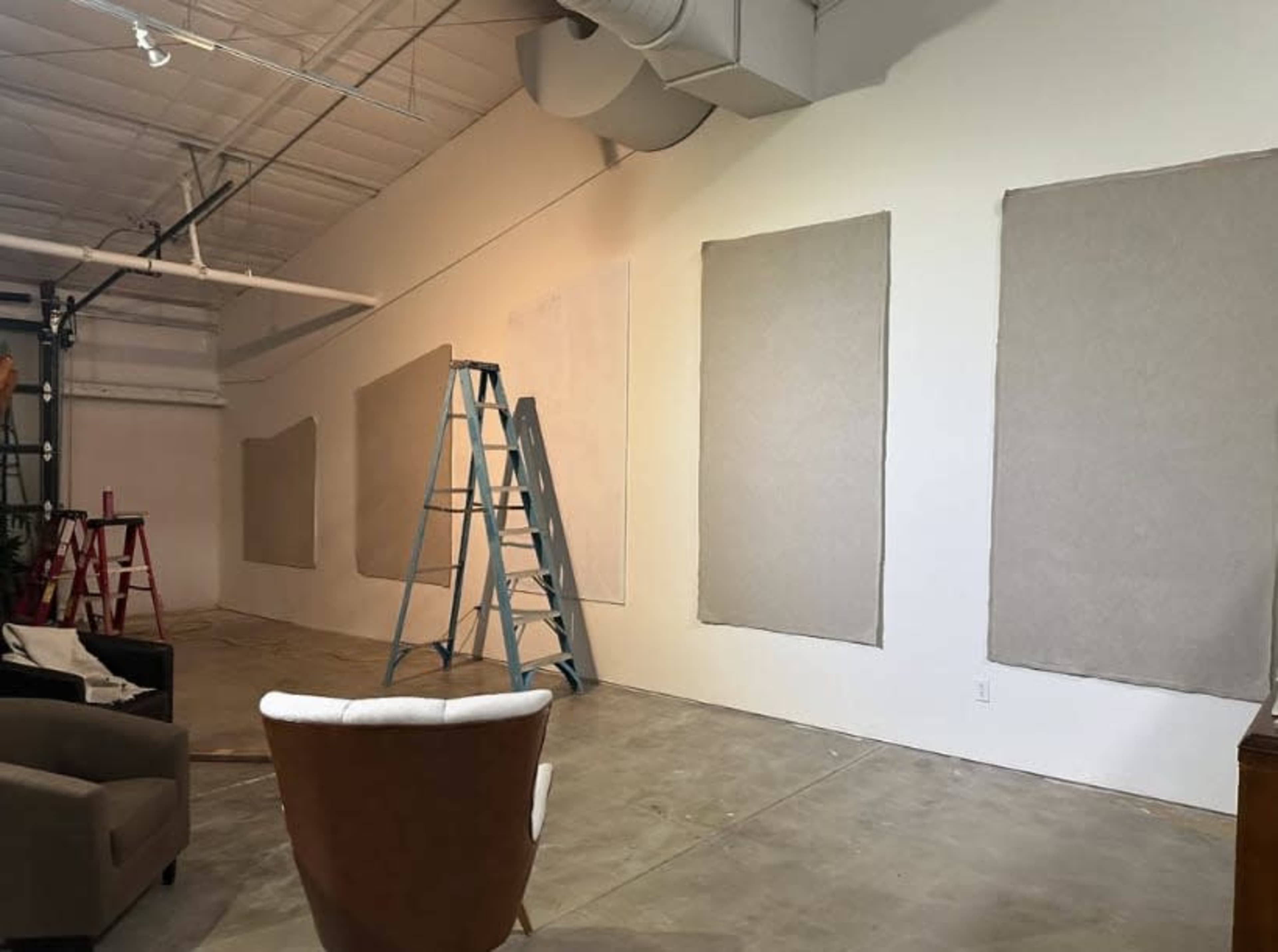 The image shows a large, empty indoor space with a bare concrete floor, white walls, and several large, unframed canvases covered in brown paper, along with a ladder positioned nearby.