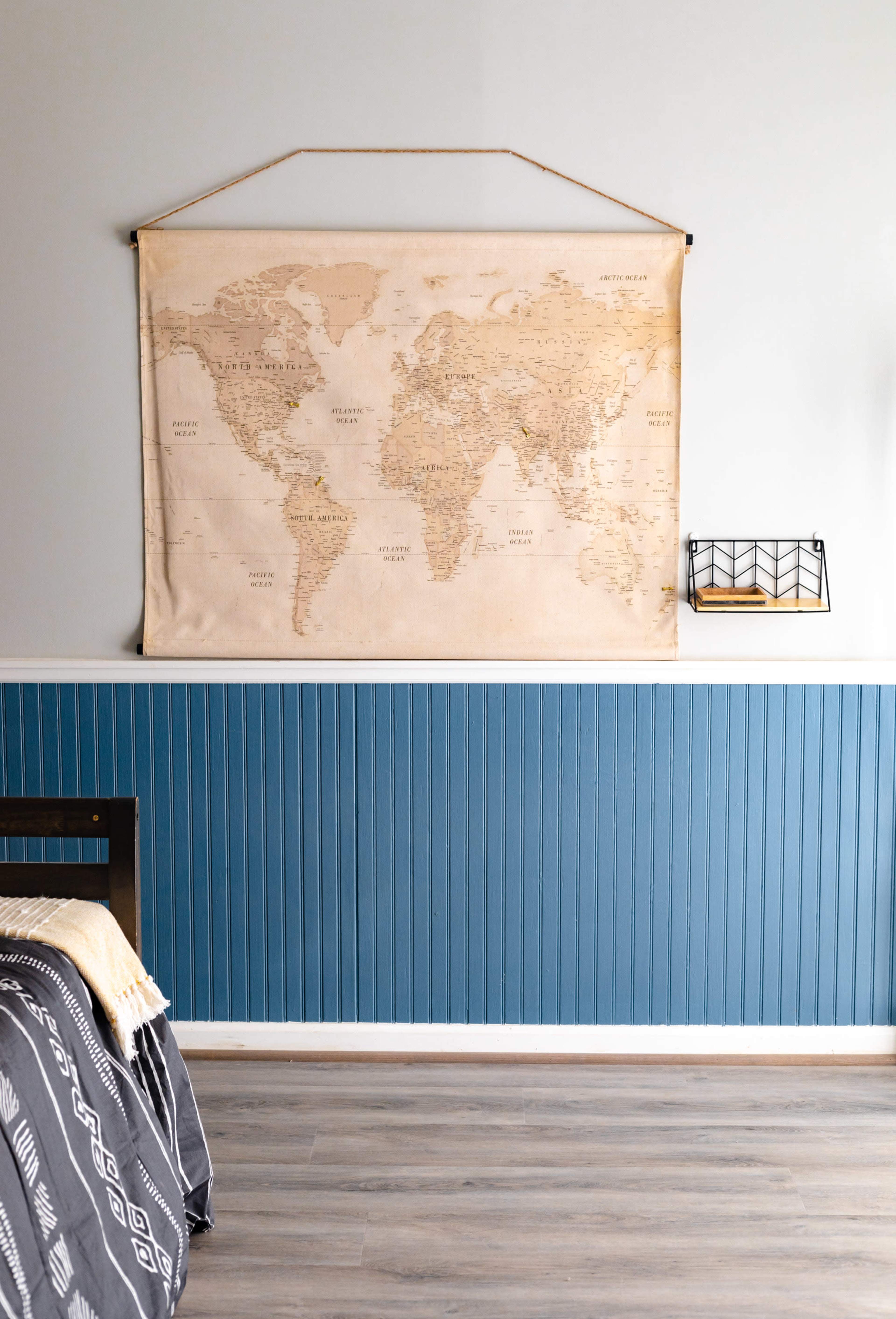 A world map hanging on a wall above a blue wainscoted section in a room with wooden flooring and a bed partially visible.